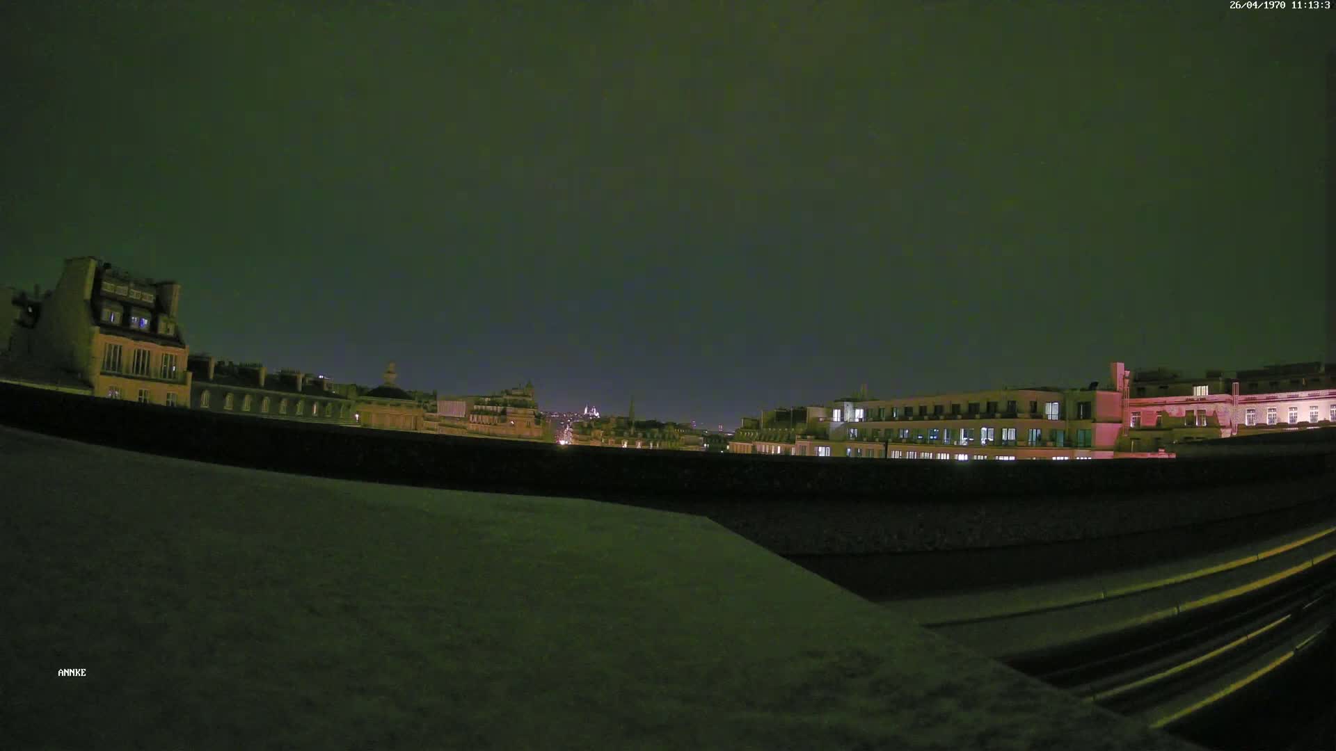 A nighttime cityscape of buildings with illuminated windows is viewed from a rooftop, under a dark, possibly overcast sky.