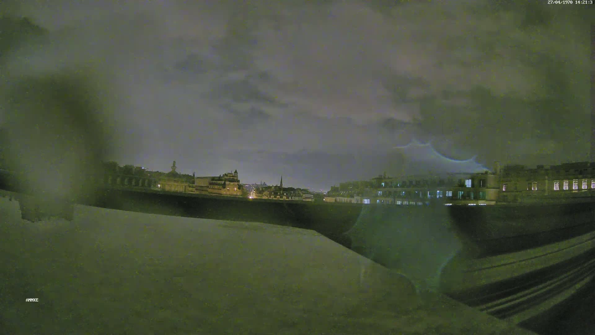 A nighttime view of a city skyline under a cloudy sky, partially obscured by rain or condensation on a window.
