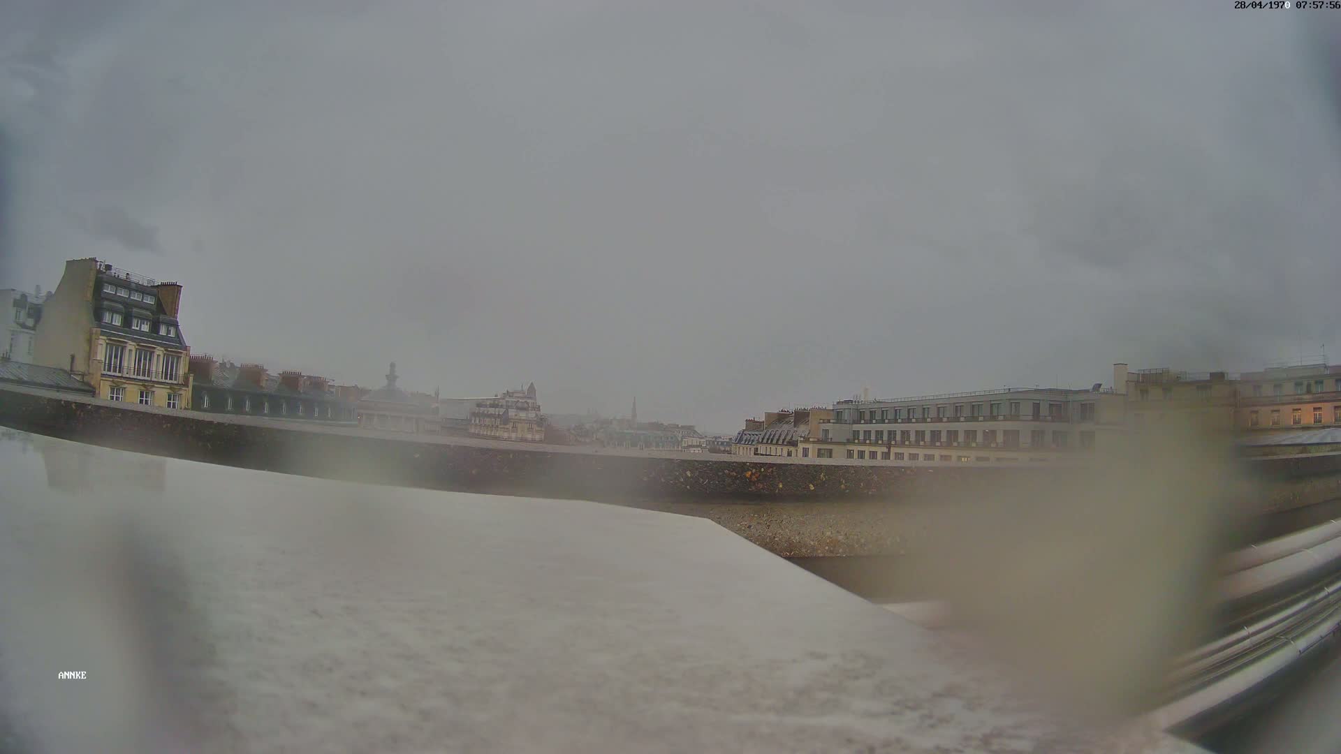 A blurry view of city buildings under an overcast sky, seen from a perspective near ground level with what appears to be rain or moisture obscuring the view.