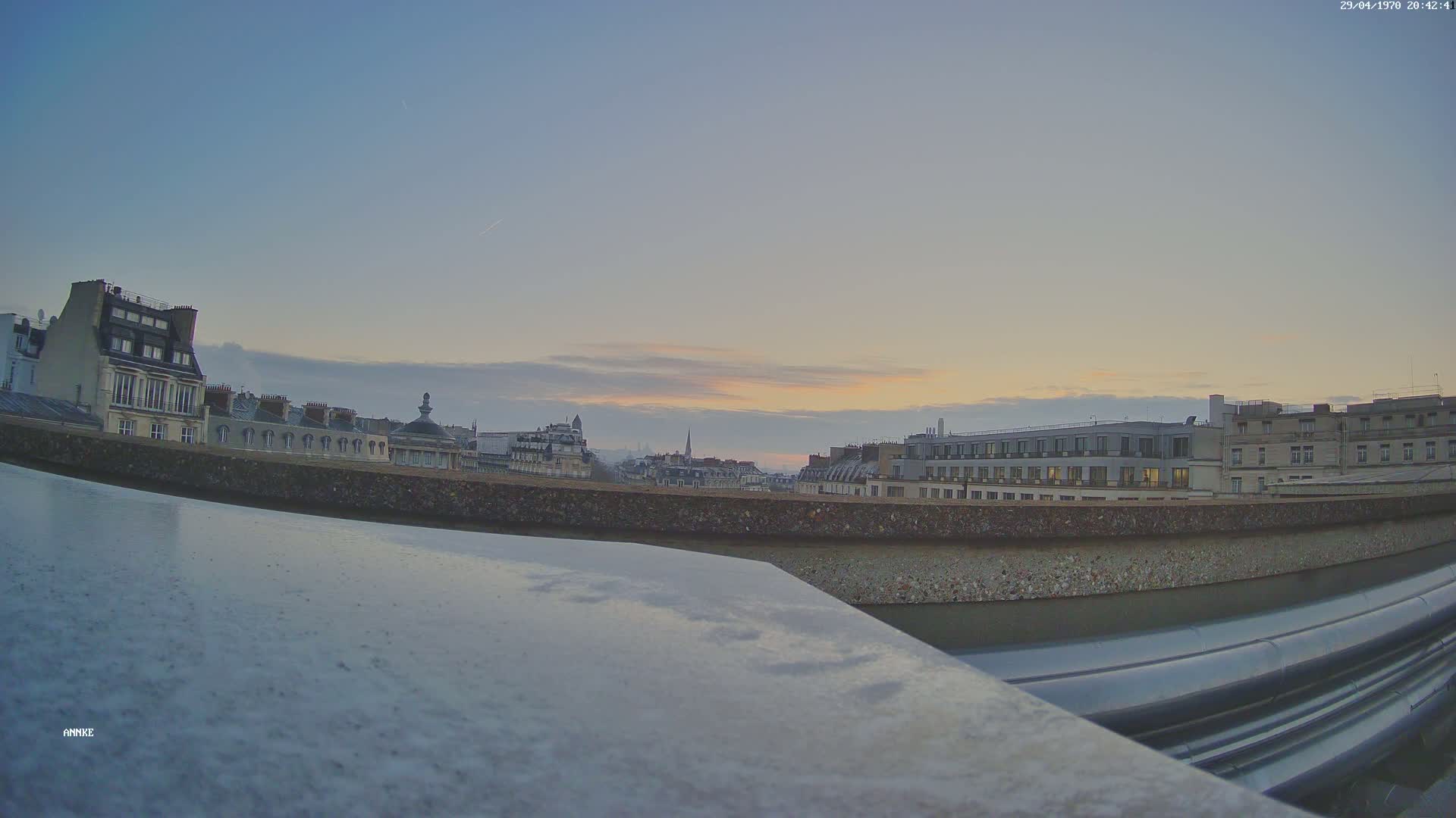 A city skyline at sunset is viewed from a rooftop, with a low-lying wall and metallic pipes in the foreground.
