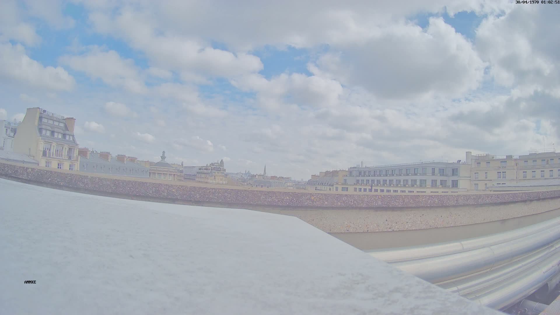 A mostly cloudy sky overlooks a cityscape of Parisian buildings, viewed from a high vantage point behind a low concrete wall and large metal pipes.