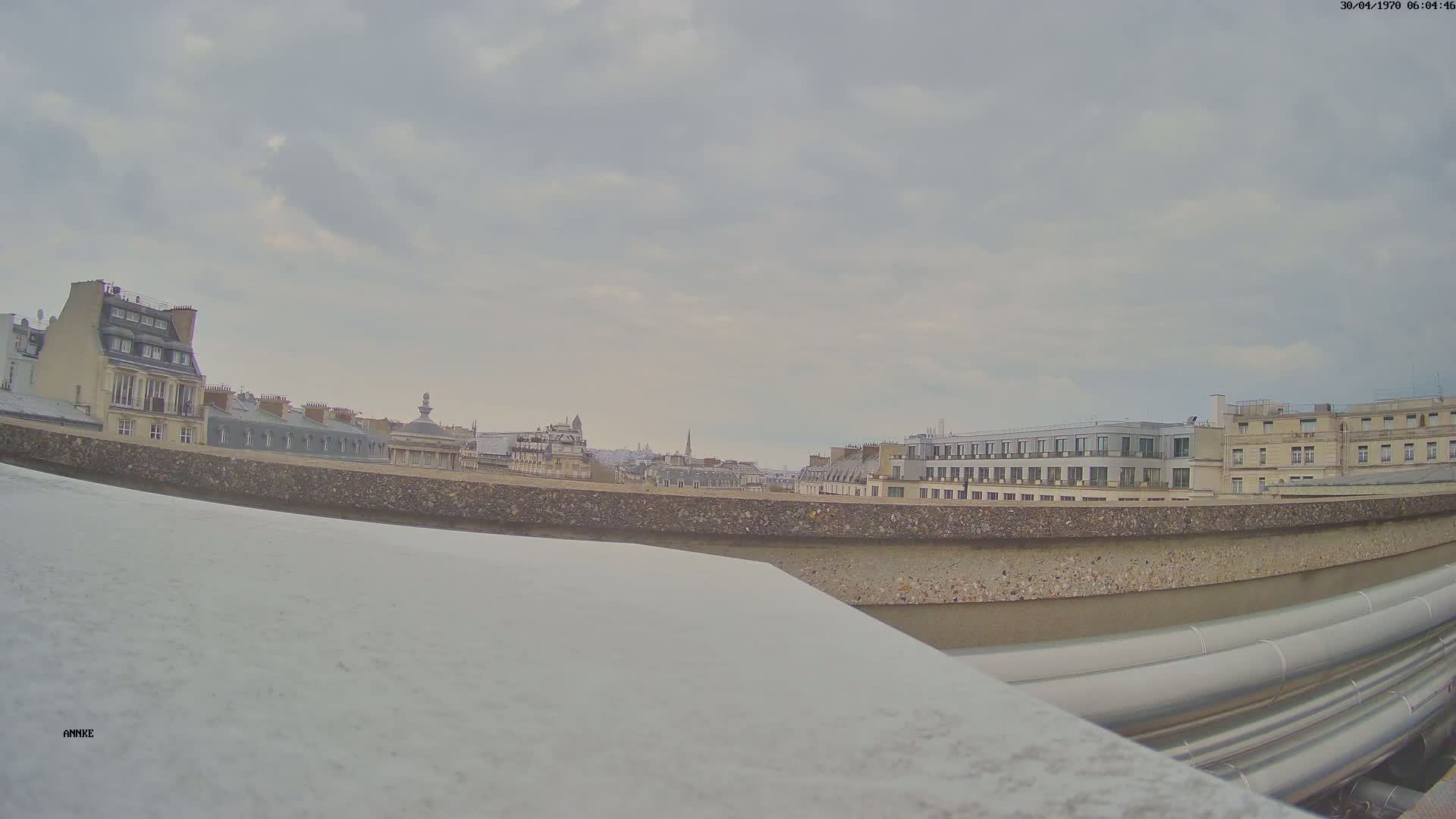 A cityscape of Paris rooftops under a cloudy sky is viewed from a high vantage point, showing large pipes in the foreground.