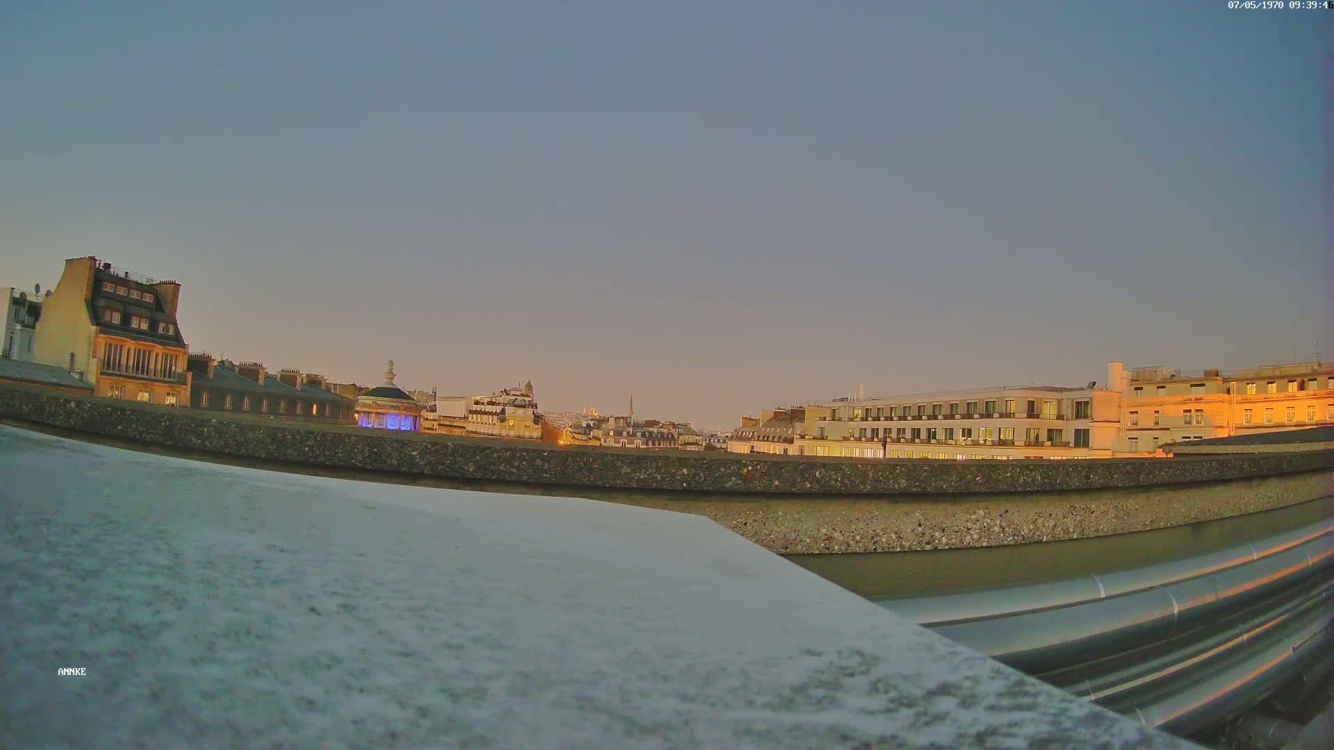 A rooftop view of city buildings at twilight under a clear sky shows a row of buildings with lit windows and a few prominent structures.
