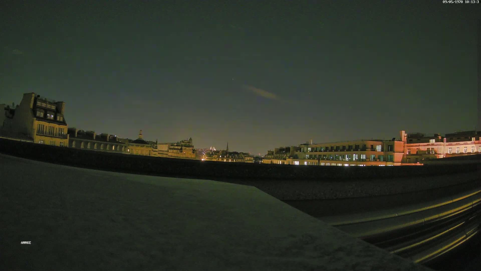 A nighttime cityscape view shows illuminated buildings under a dark, mostly clear sky, seen from a rooftop vantage point.