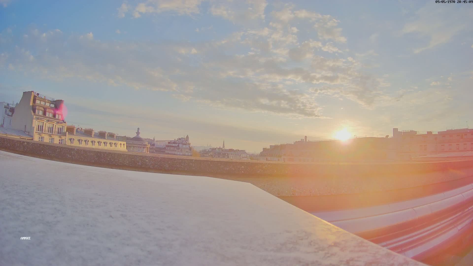 A city skyline at sunset is viewed from a rooftop, with a bright sun partially obscured by lens flare.