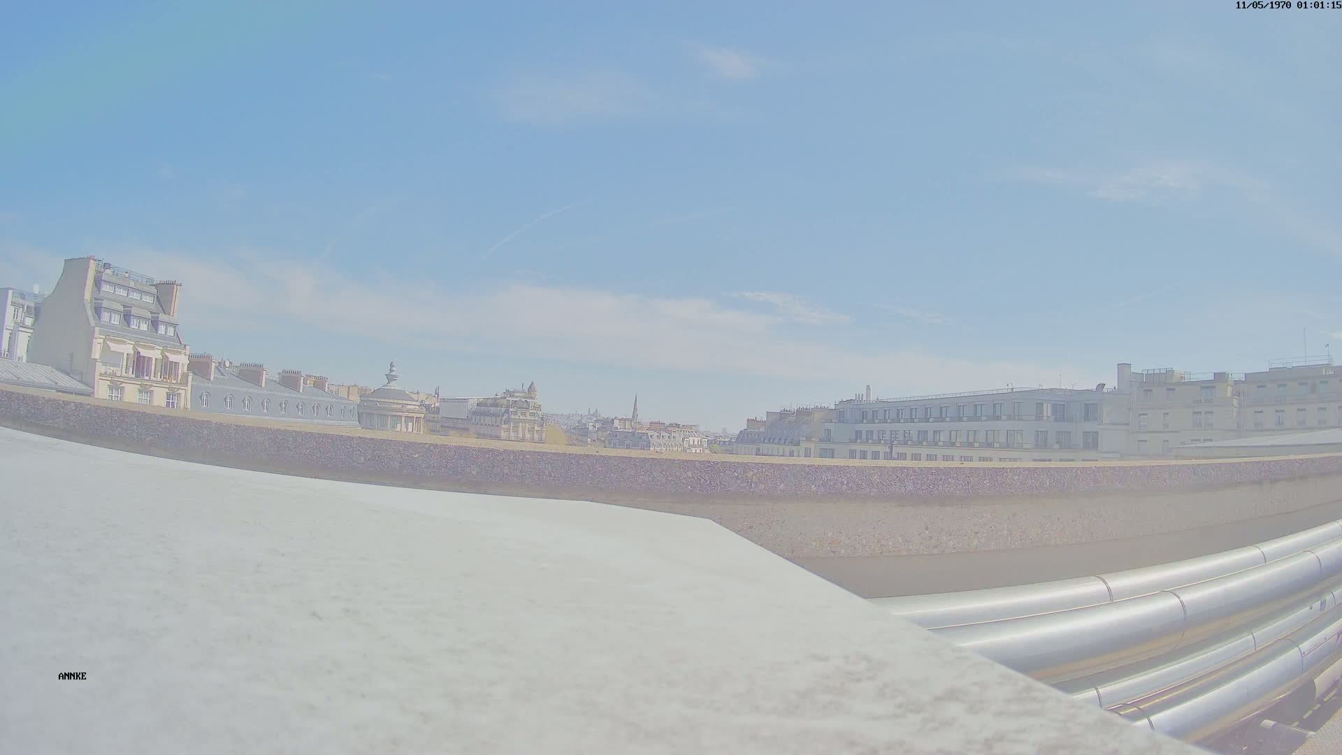 A cityscape of Parisian buildings under a mostly clear, bright blue sky is viewed from a rooftop, with large industrial pipes in the foreground.