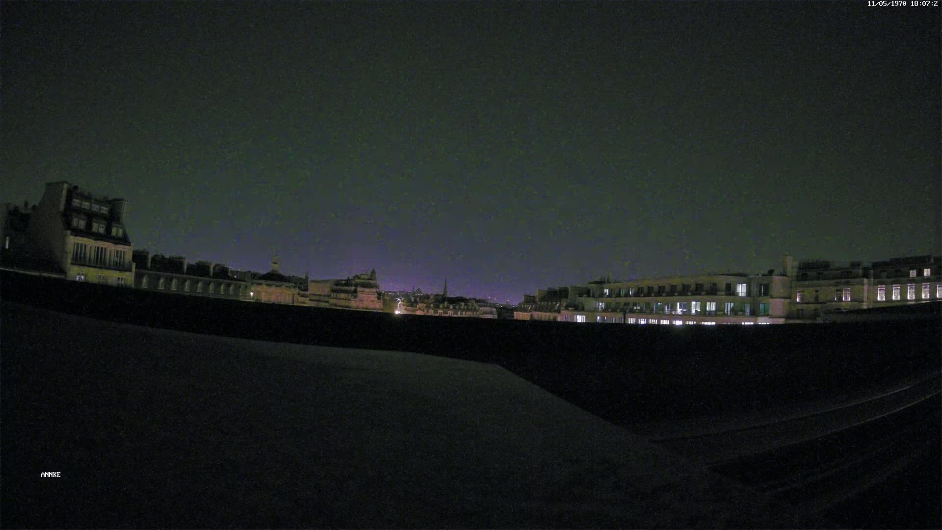 A nighttime view from a rooftop shows a cityscape of dark buildings with some illuminated windows under a dark, clear sky.