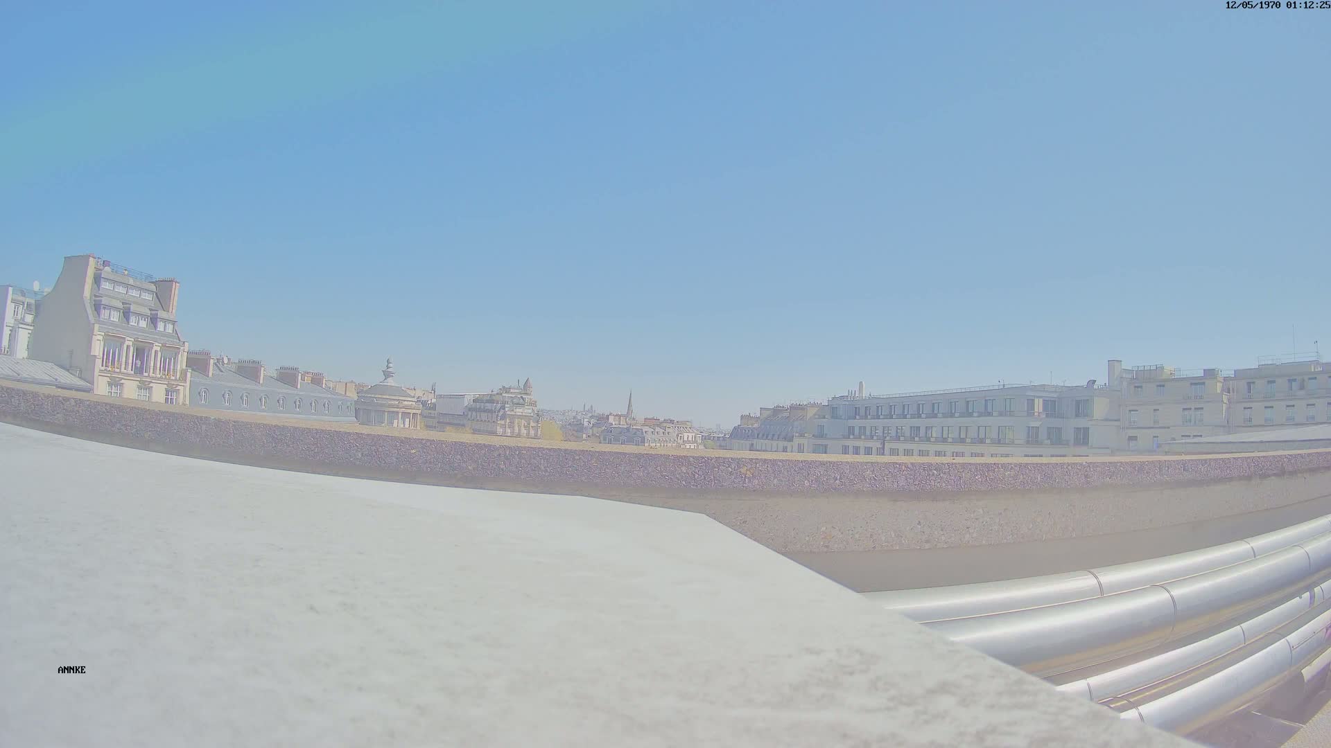 A cityscape viewed from a rooftop on a clear, sunny day, showing several buildings and large metallic pipes in the foreground.