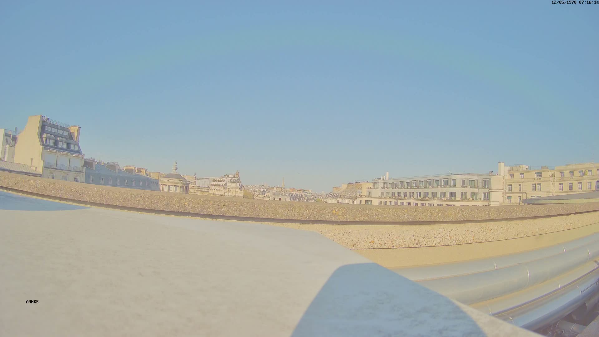 A low-angle view from a rooftop shows a cityscape under a clear blue sky, with several multi-story buildings visible in the distance.