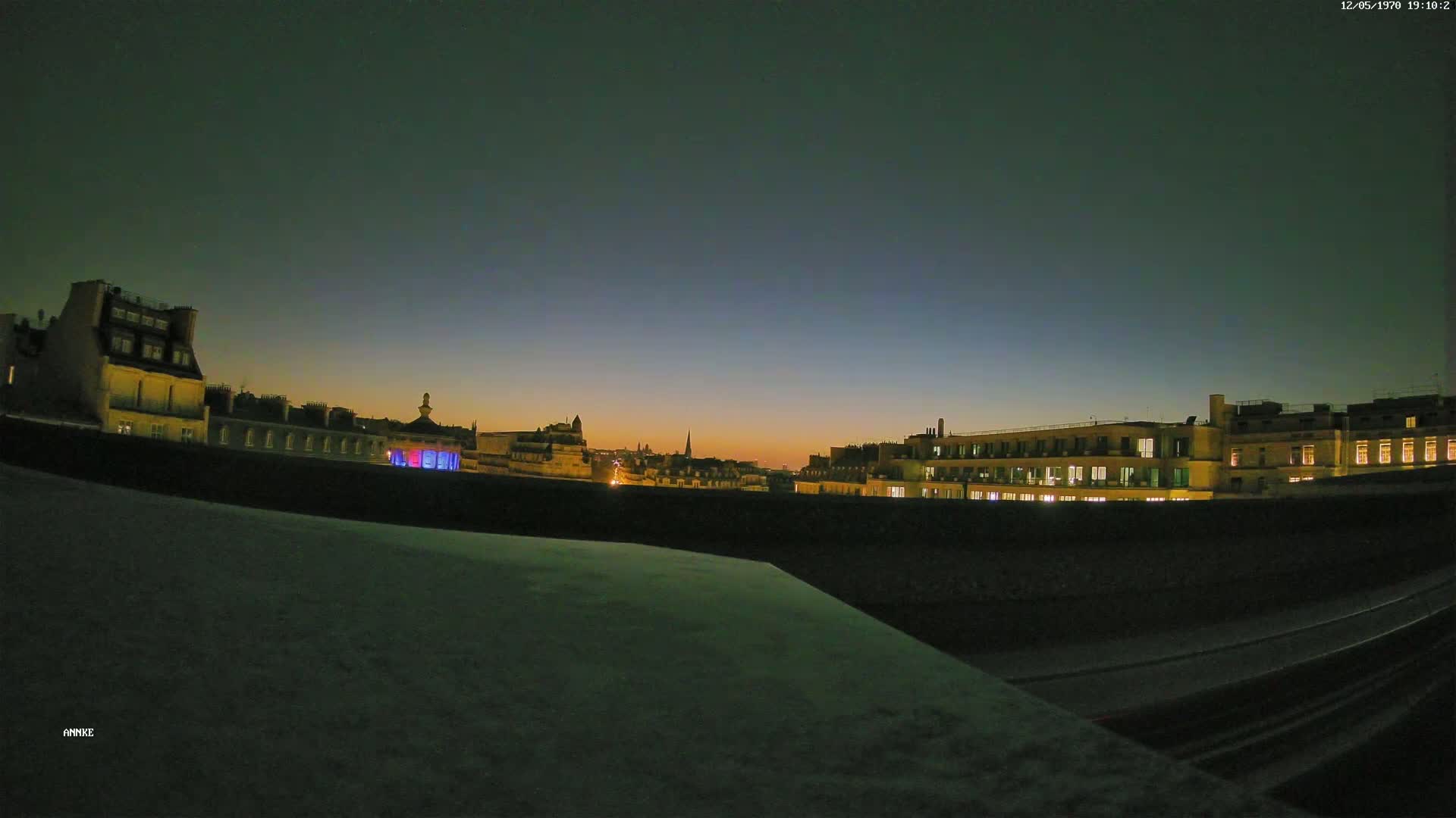 A nighttime cityscape with illuminated buildings and a dark, clear sky is seen from a high vantage point.