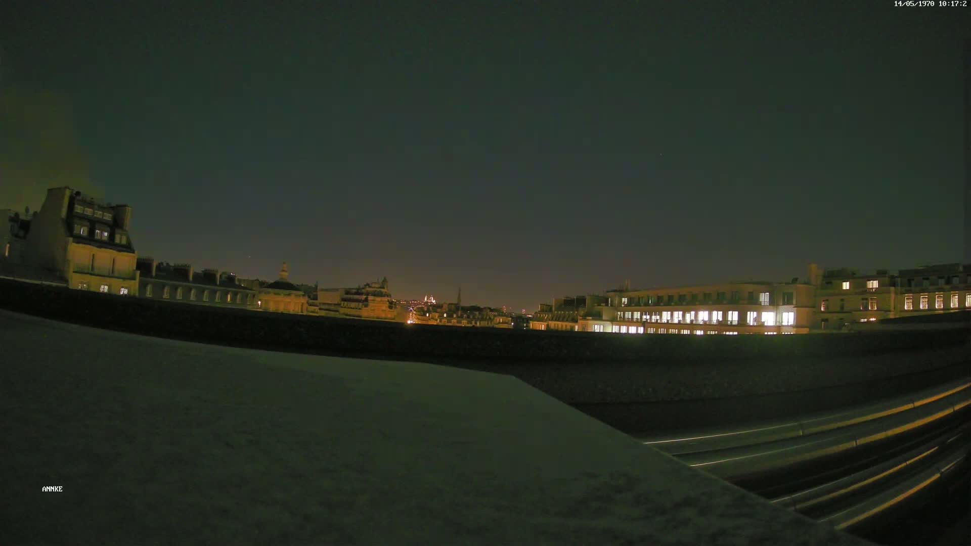 A nighttime cityscape of illuminated buildings and a roadway with light trails under a dark sky.