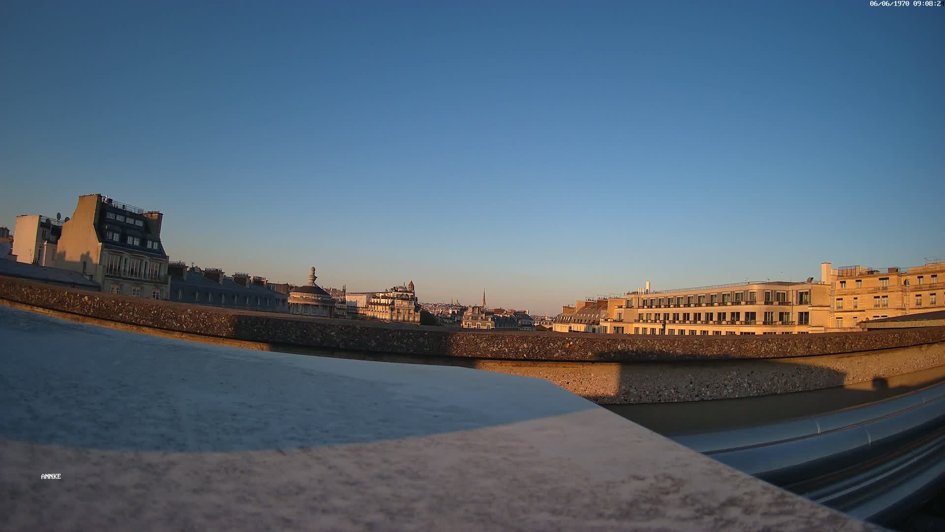 A cityscape of Parisian buildings at sunset under a clear blue sky is viewed from a rooftop, partially obscured by a low wall.