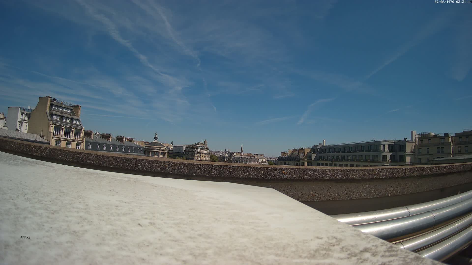 A sunny cityscape view from a rooftop shows a row of buildings under a mostly clear blue sky, with large silver pipes in the foreground.