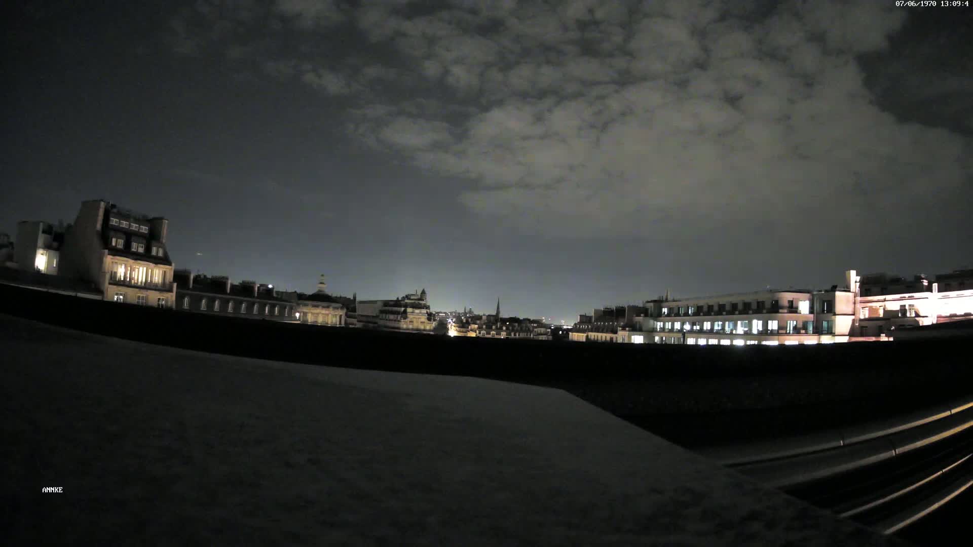 A nighttime cityscape with a cloudy sky is viewed from a rooftop, showing illuminated buildings and a dark, snow-covered area in the foreground.
