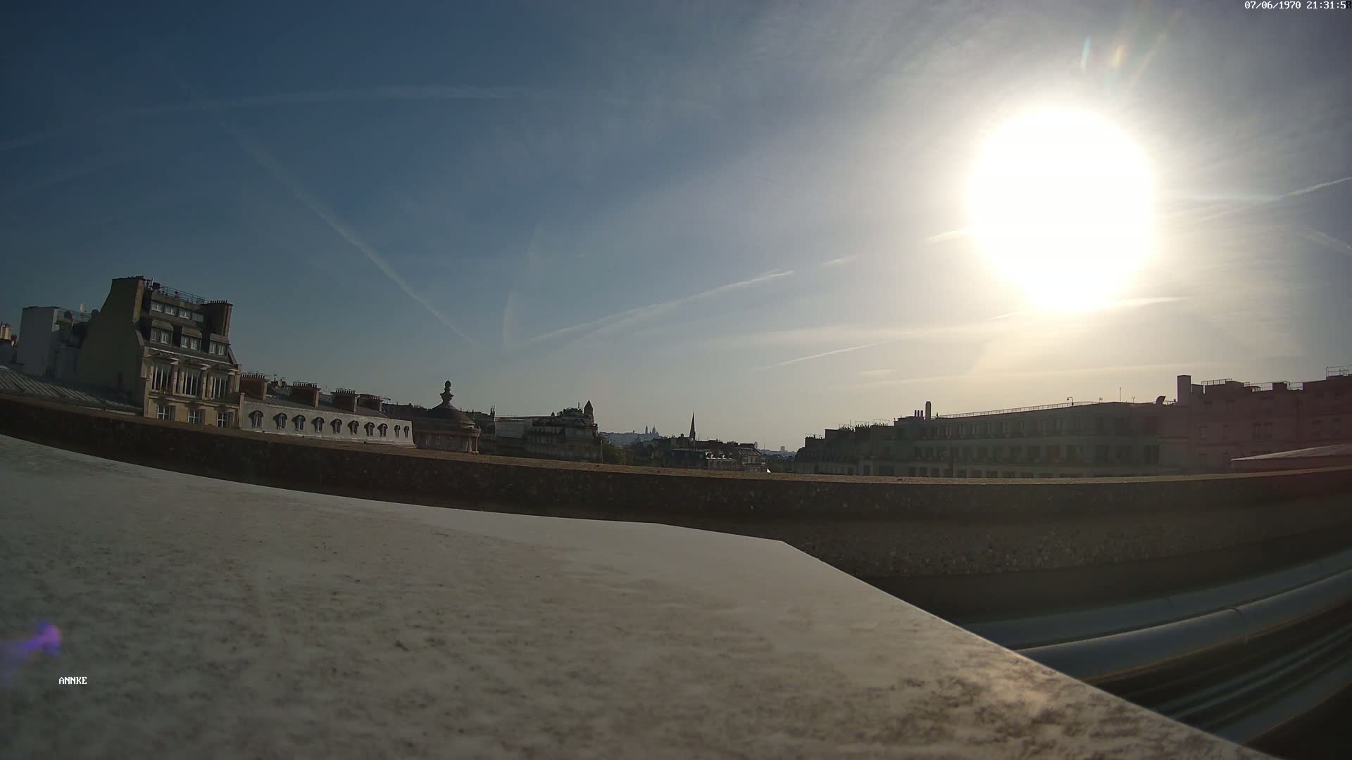 A sunny cityscape is viewed from a rooftop, showing a row of buildings and a distant skyline.