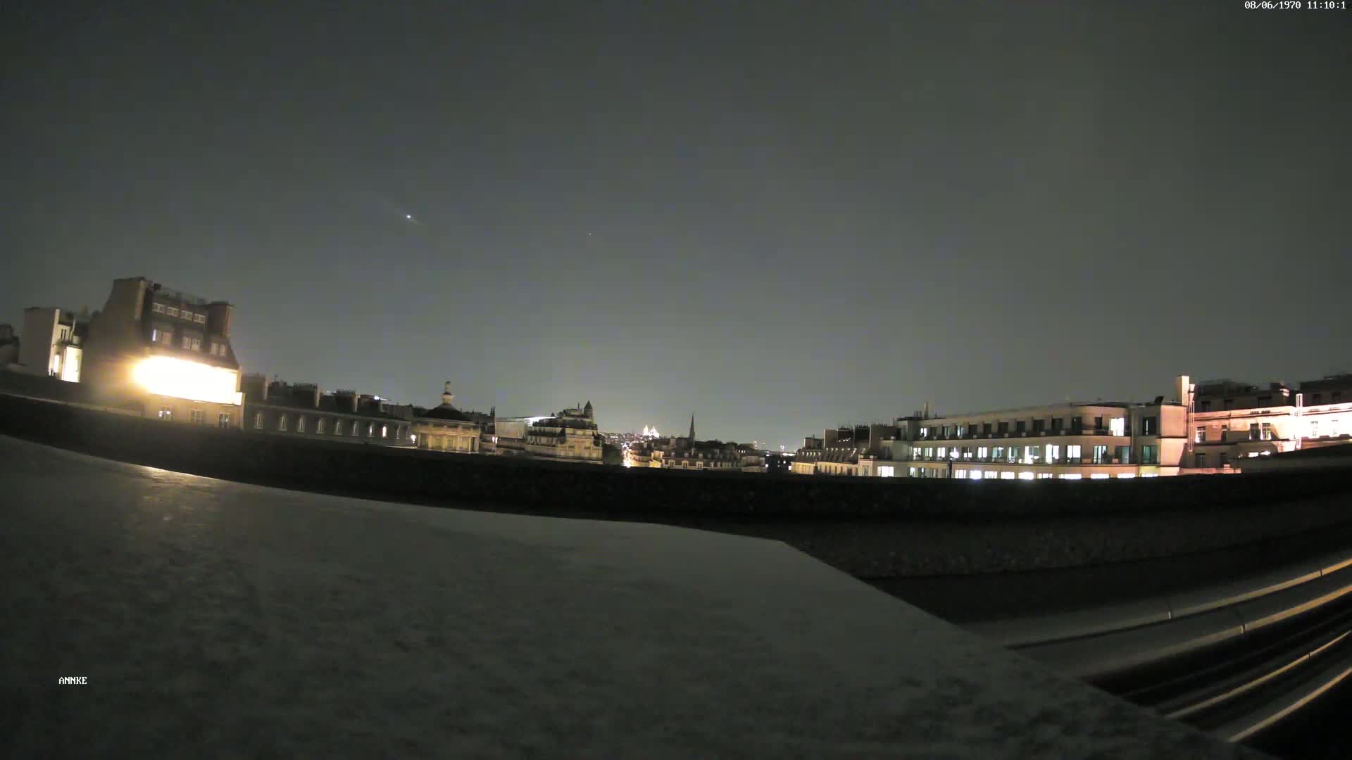A nighttime city skyline is visible from a rooftop, under a dark, clear sky.