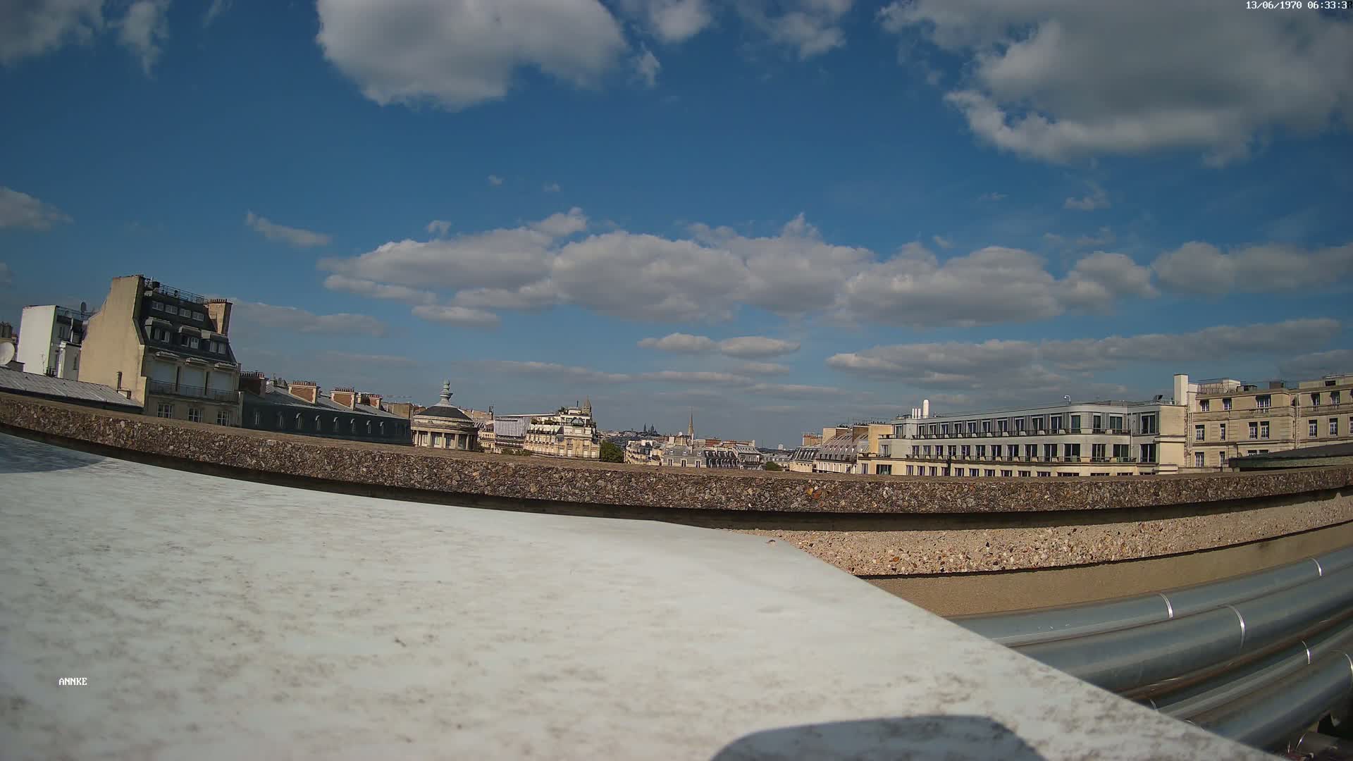 A cityscape view from a rooftop shows a partly cloudy blue sky, with several multi-story buildings in the background.