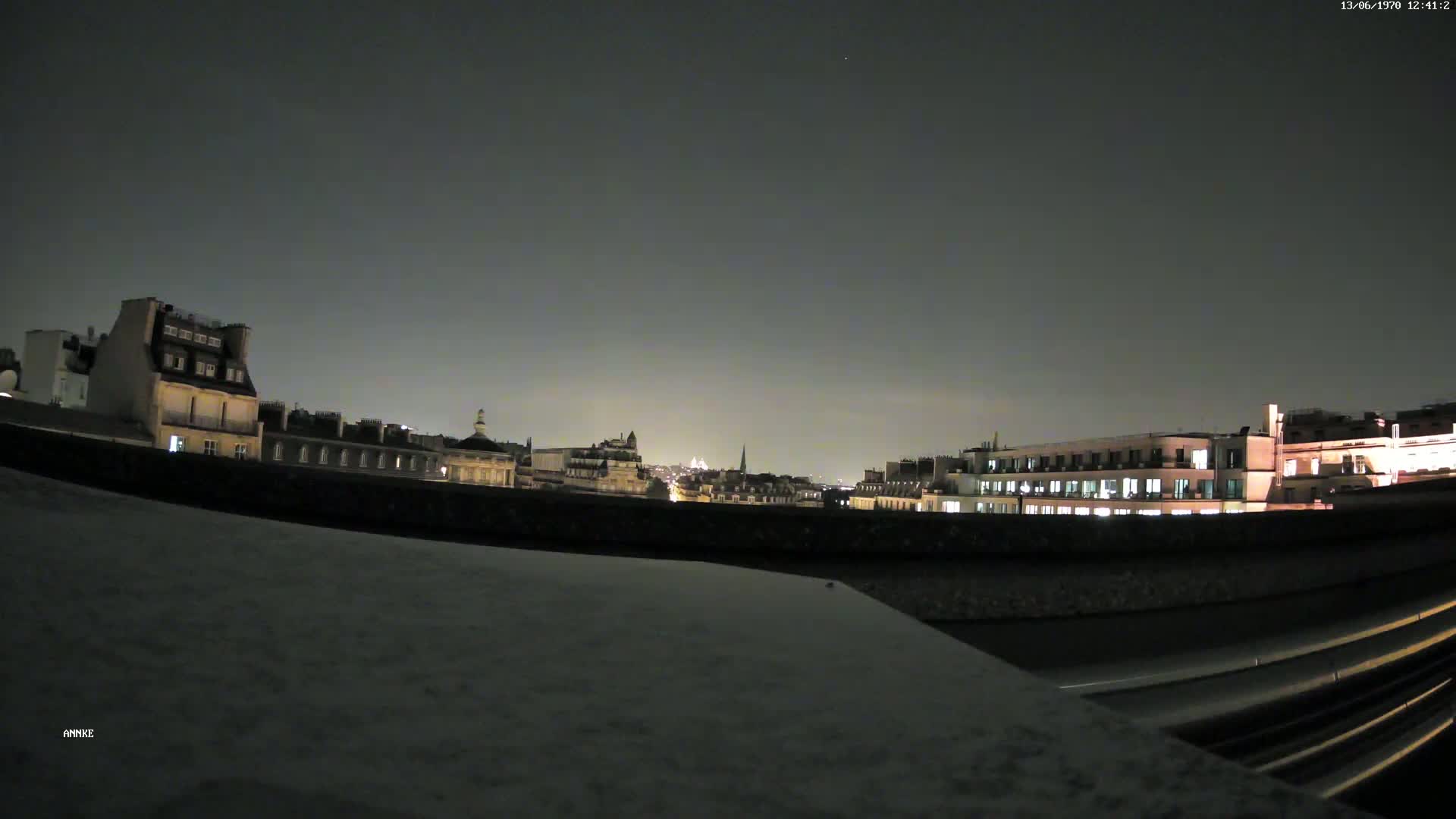 A nighttime cityscape of Parisian buildings is visible from a rooftop, under a dark, clear sky.