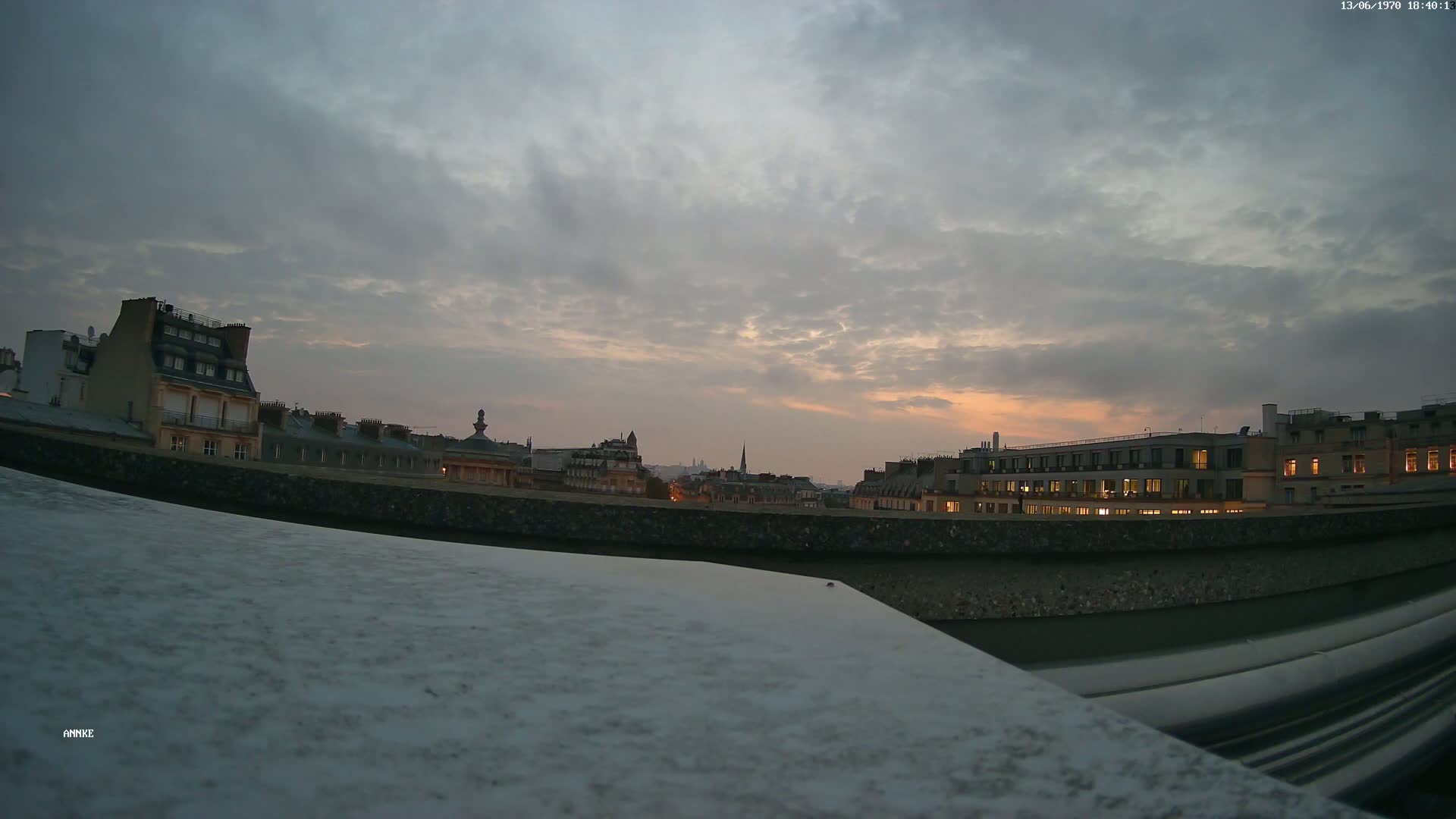 A city skyline at dusk under a cloudy sky is visible from a high vantage point above a flat, light-grey surface.