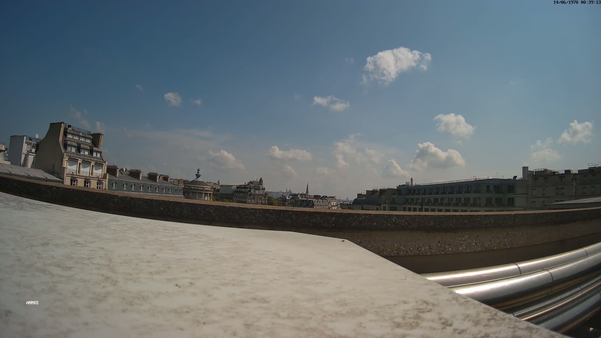 A rooftop view of a city on a partly sunny day shows several buildings, a low wall, and large metallic pipes.