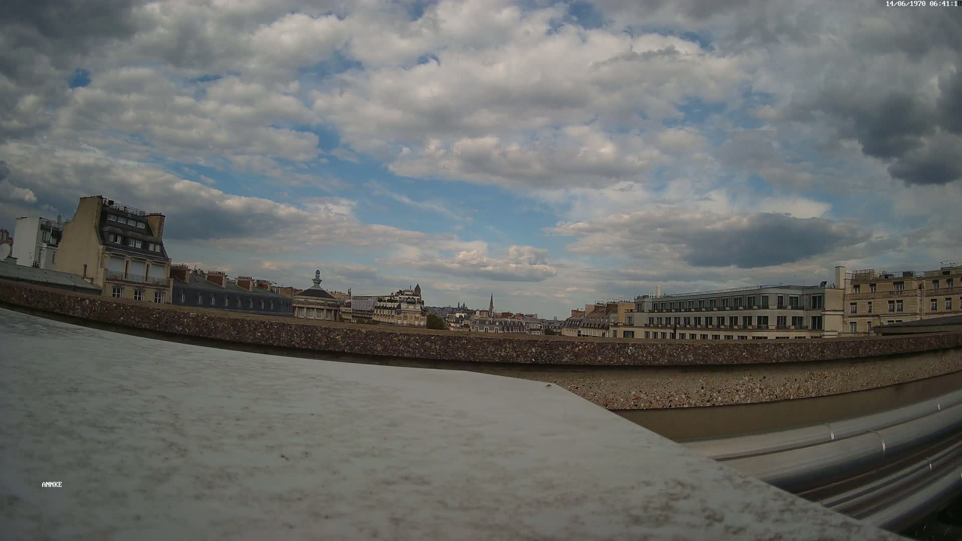 A partly cloudy sky overlooks a cityscape of Parisian-style buildings viewed from a rooftop, with a low wall and metallic pipes in the foreground.