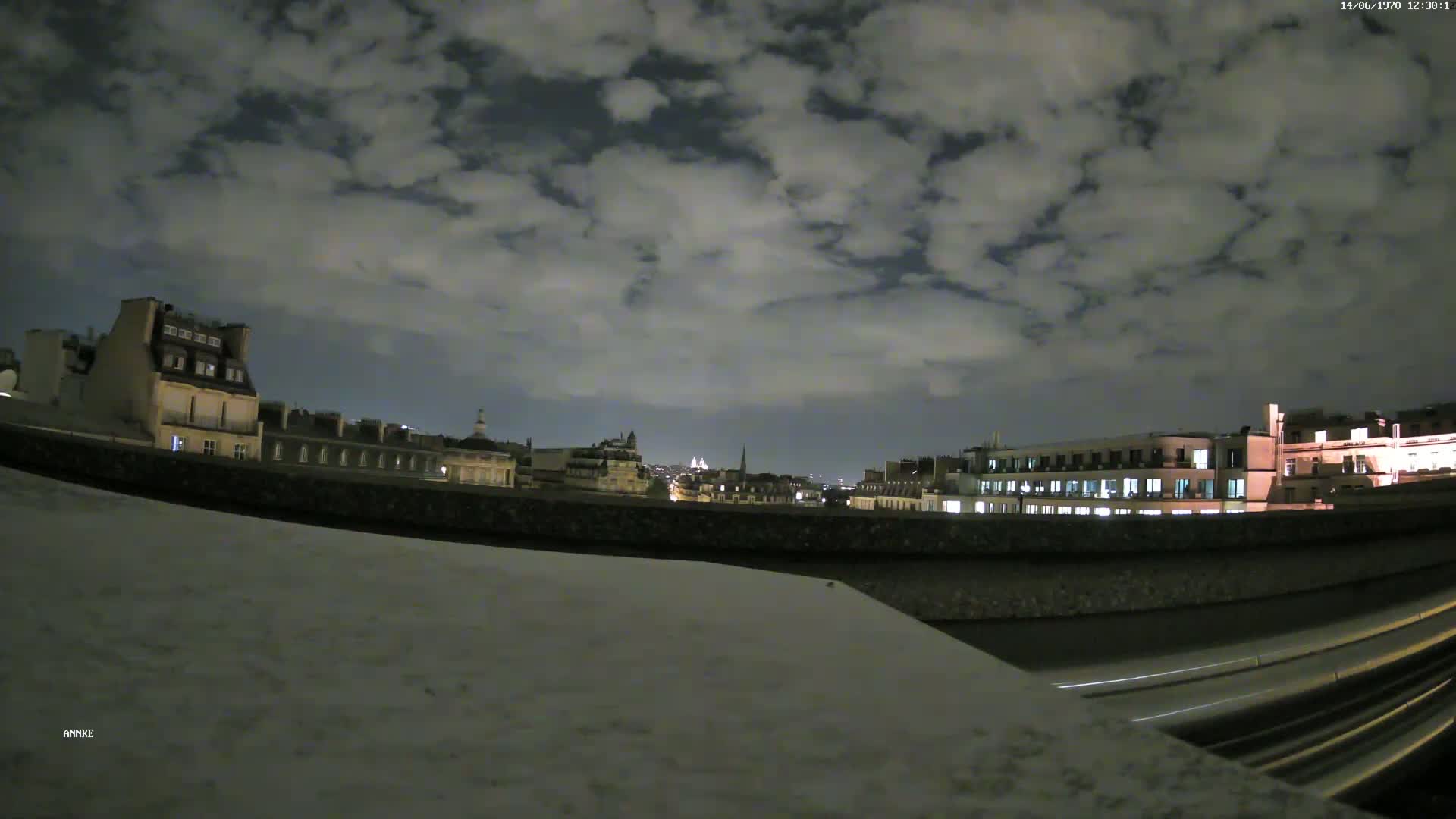 A nighttime view of a city skyline with a cloudy sky, seen from a rooftop overlooking a low wall and a road below.