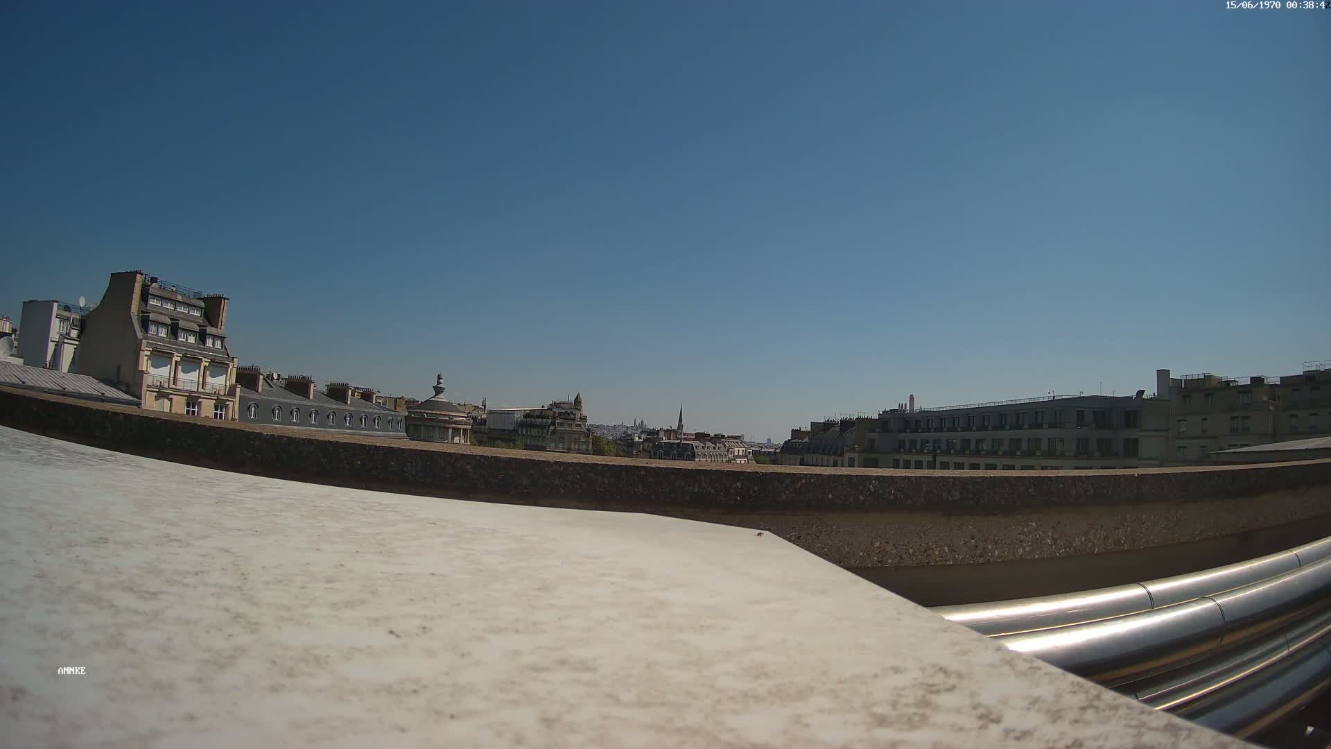 A rooftop view on a clear, sunny day shows a cityscape of Parisian-style buildings, with large metallic pipes in the foreground.