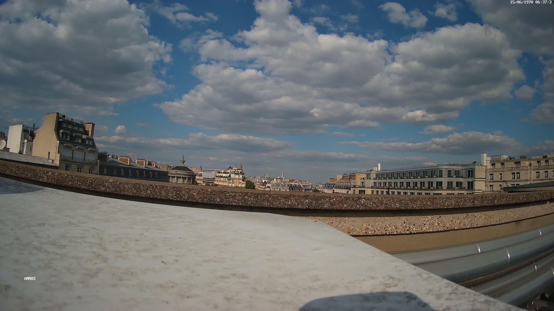 A partly cloudy sky overlooks a cityscape of Parisian buildings viewed from a rooftop, with large metal pipes visible in the foreground.
