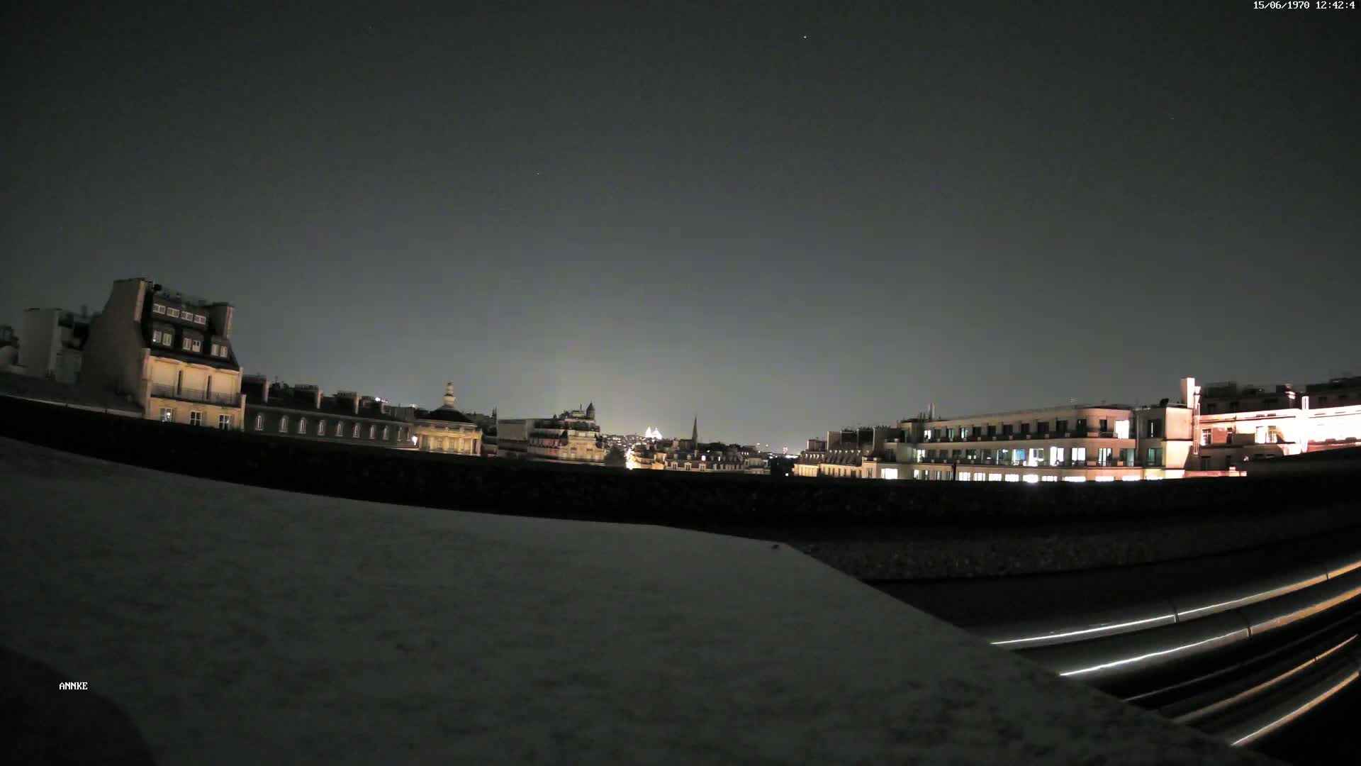 A nighttime cityscape of illuminated buildings is visible from a rooftop, under a dark, clear sky.