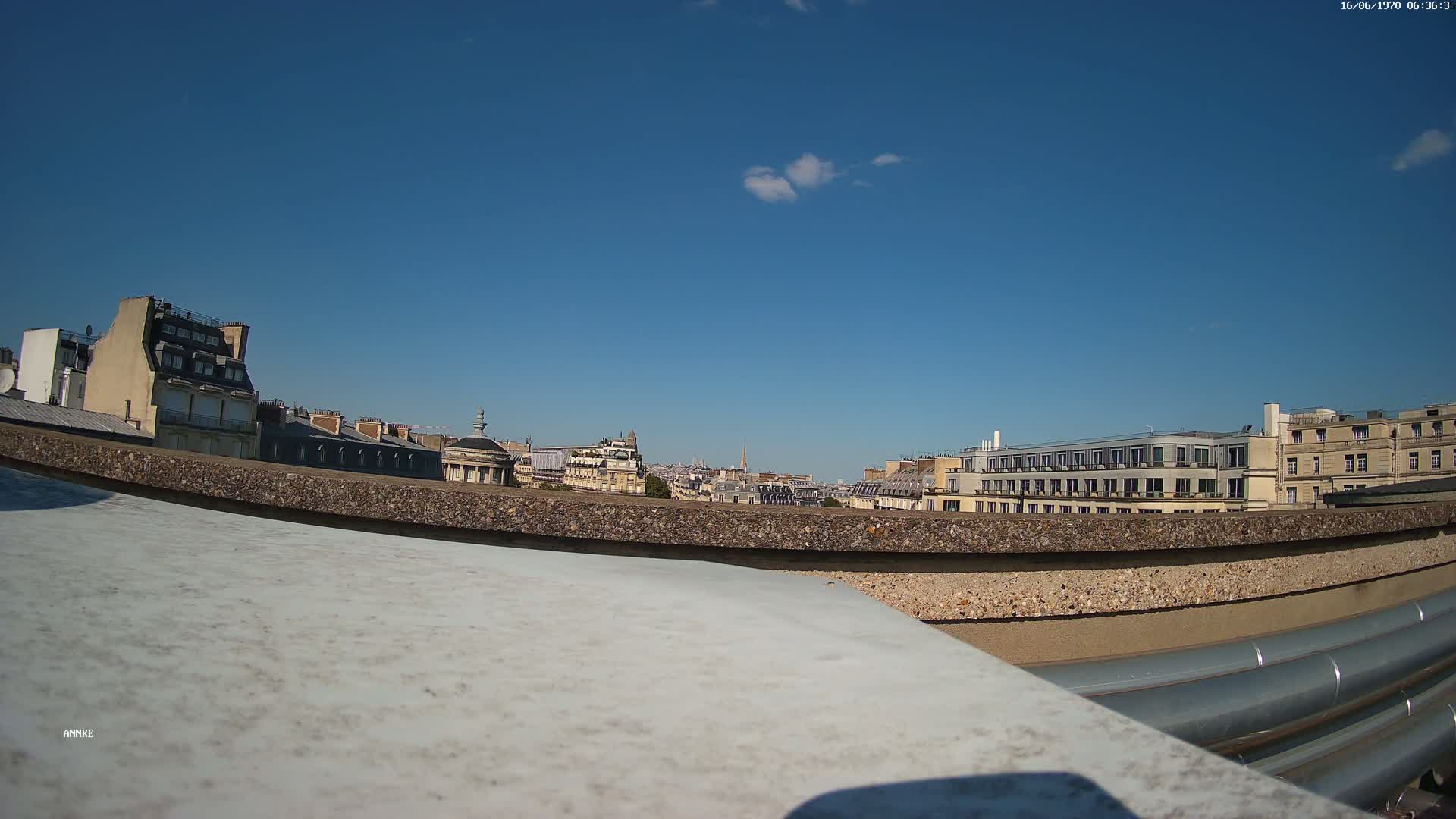 A clear blue sky overlooks a city skyline of Parisian-style buildings viewed from a rooftop, with large metal pipes in the foreground.