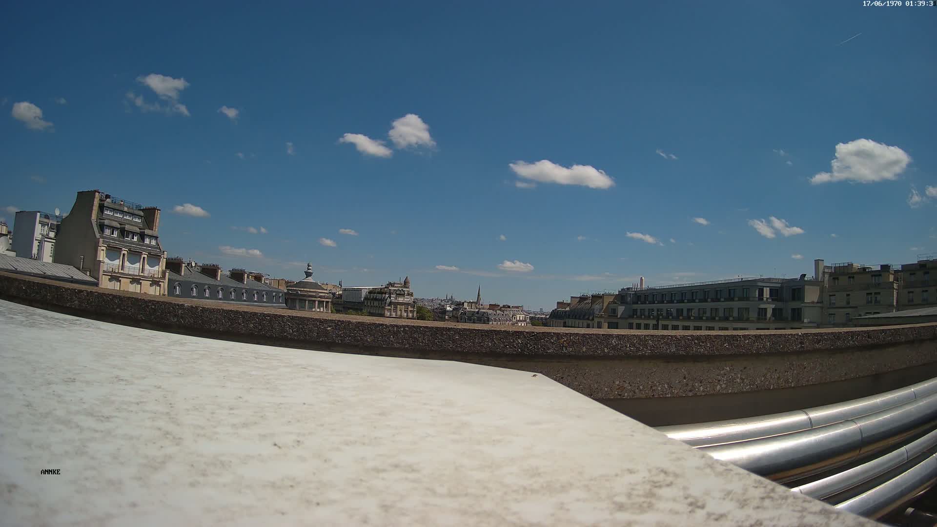 A rooftop view shows a city skyline under a mostly sunny sky with a few scattered clouds, and large metal pipes in the foreground.