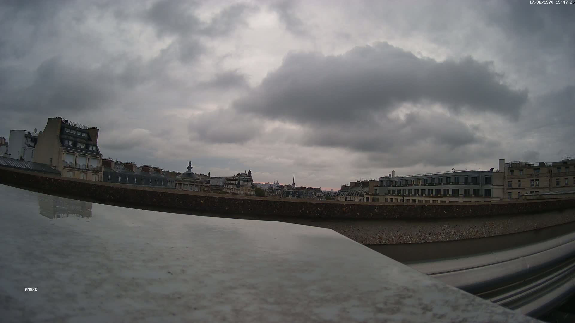 A cityscape of Parisian buildings is visible under a cloudy, overcast sky.
