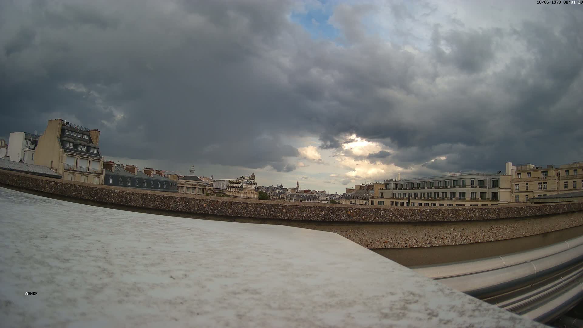 A cityscape of Parisian buildings is visible under a dark, cloudy sky, with a hint of sunlight breaking through.