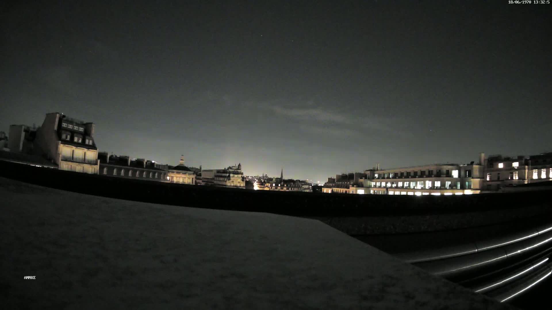 A nighttime view of a city skyline under a dark, mostly clear sky, seen from a rooftop with a curved metal structure in the foreground.