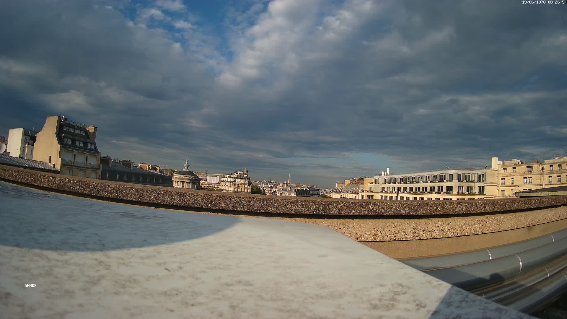 A rooftop view of a city under a partly cloudy sky shows several buildings, including a large, modern structure.