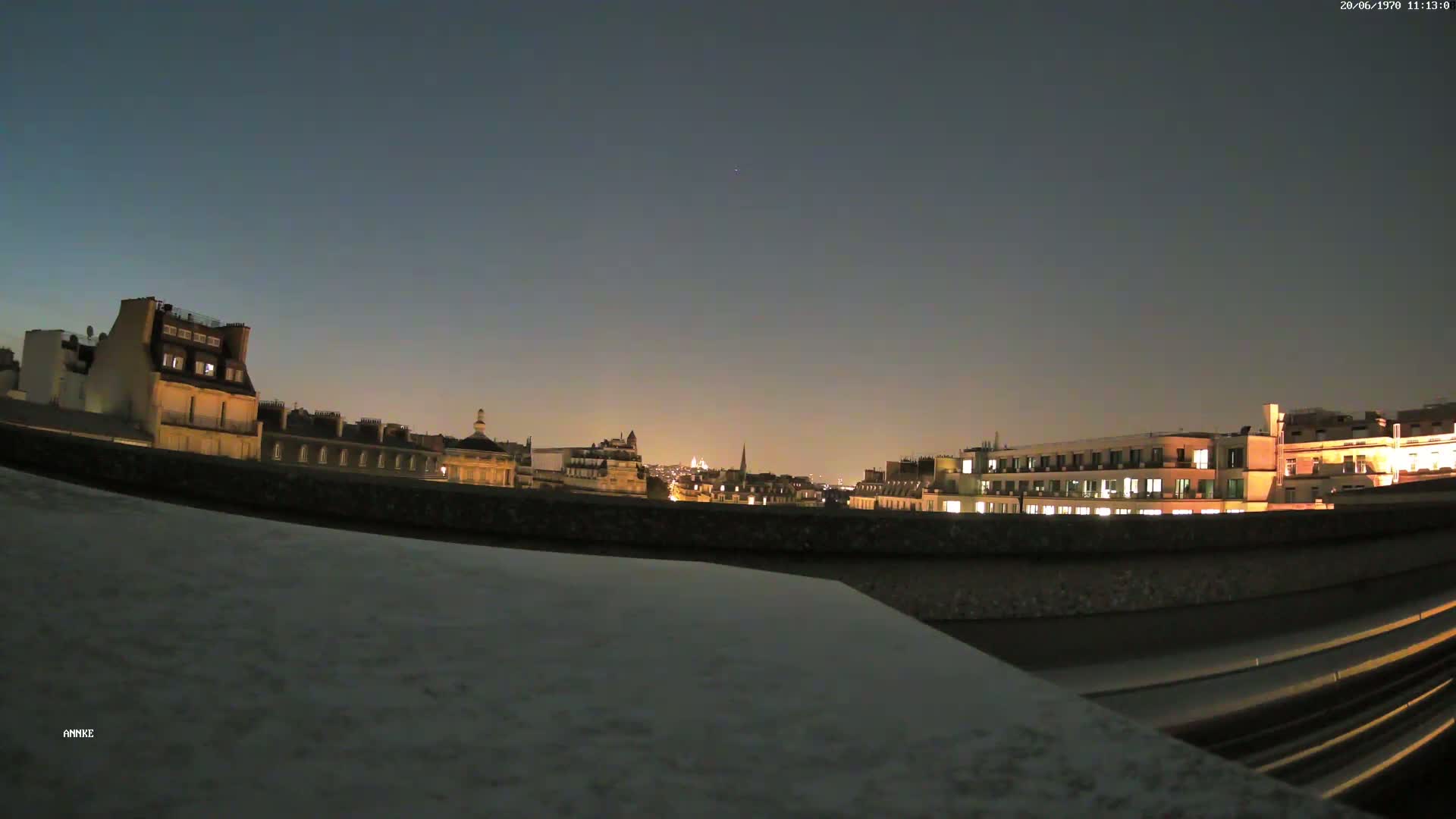 A nighttime cityscape of illuminated buildings is viewed from behind a low wall under a clear, dark sky.