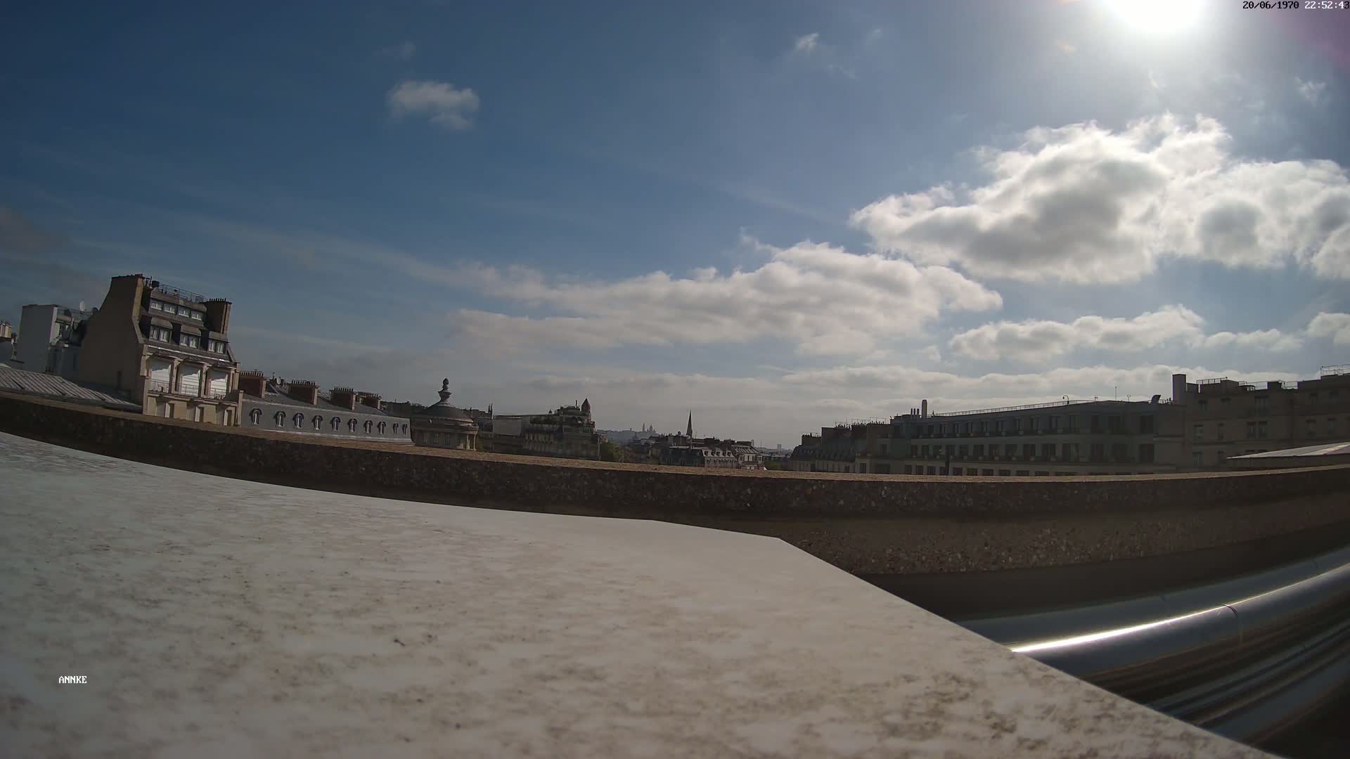 A partly sunny day reveals a cityscape of Parisian buildings viewed from a rooftop, with a low wall and metallic pipes in the foreground.