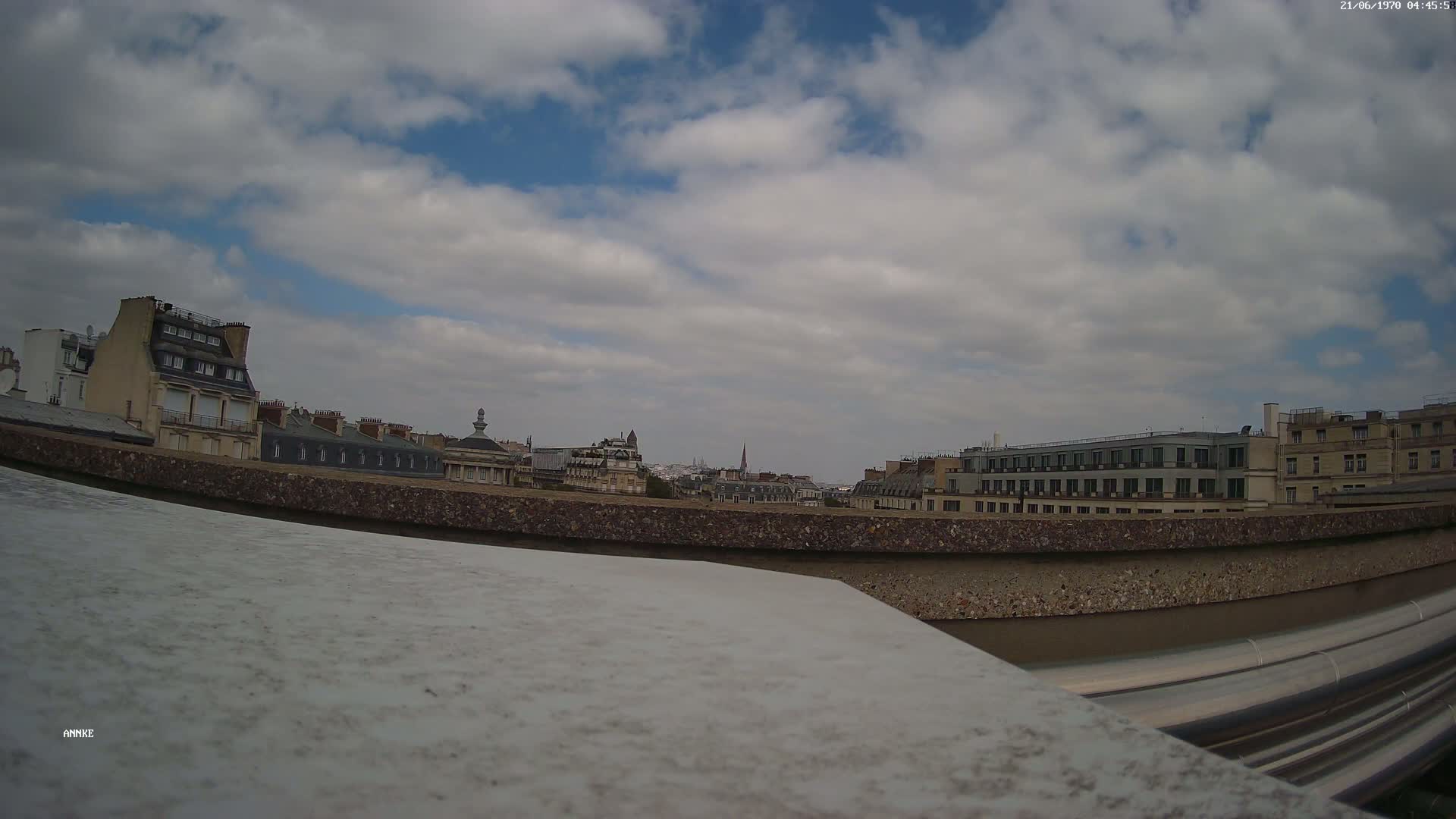 A partly cloudy sky overlooks a cityscape of Parisian buildings viewed from a rooftop, with a low-lying concrete wall and metallic pipes in the foreground.