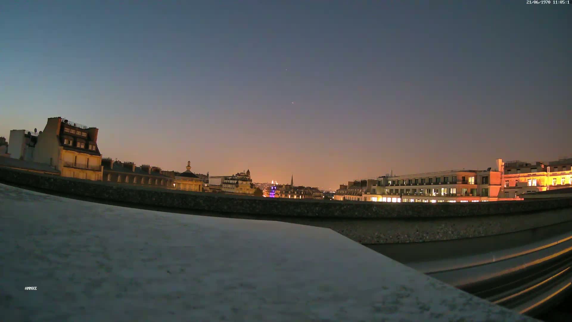 A nighttime cityscape is viewed from a rooftop, showing illuminated buildings under a twilight sky.