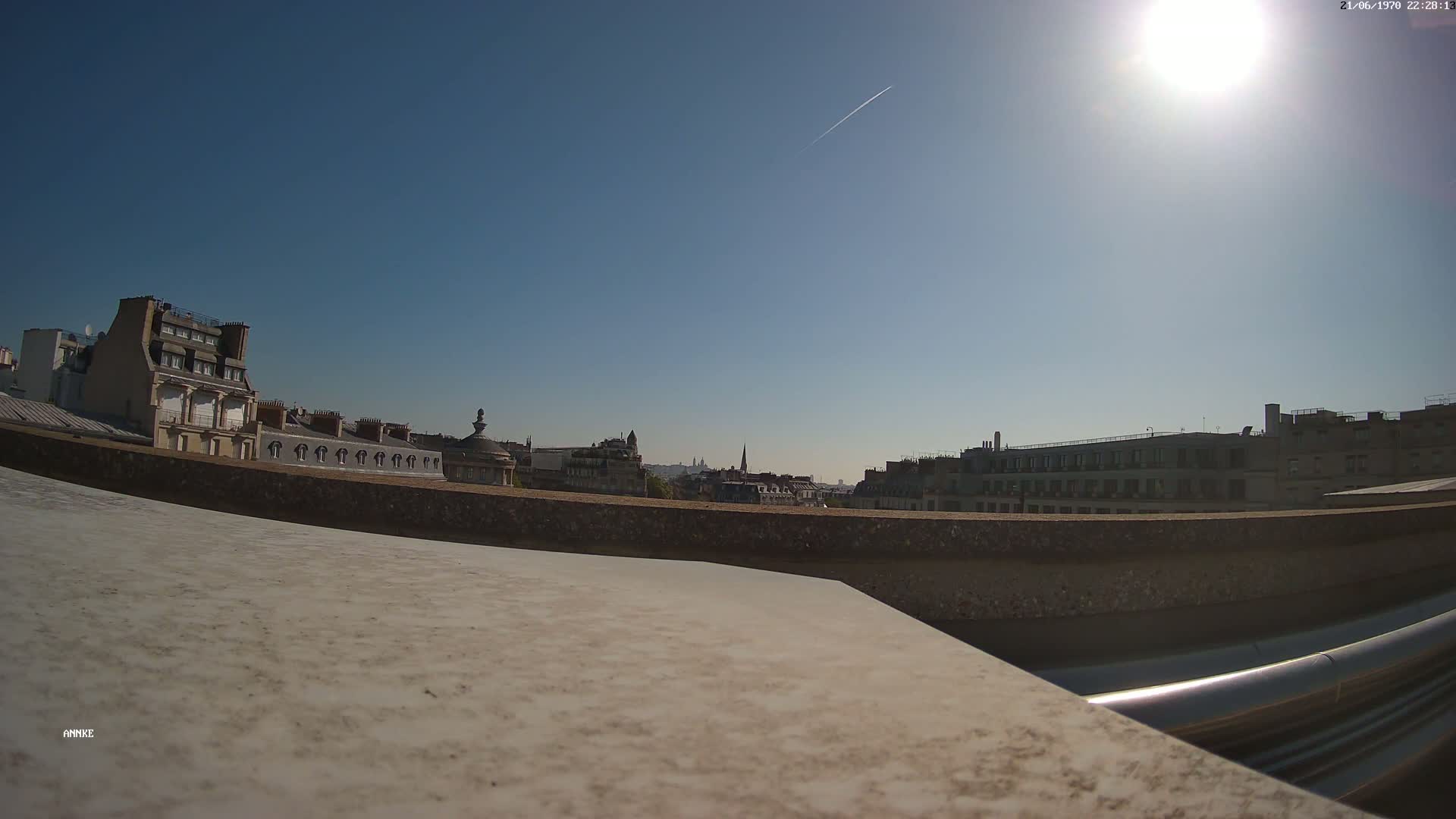 A sunny cityscape is viewed from a rooftop, showing a row of buildings and a clear blue sky with a contrail.