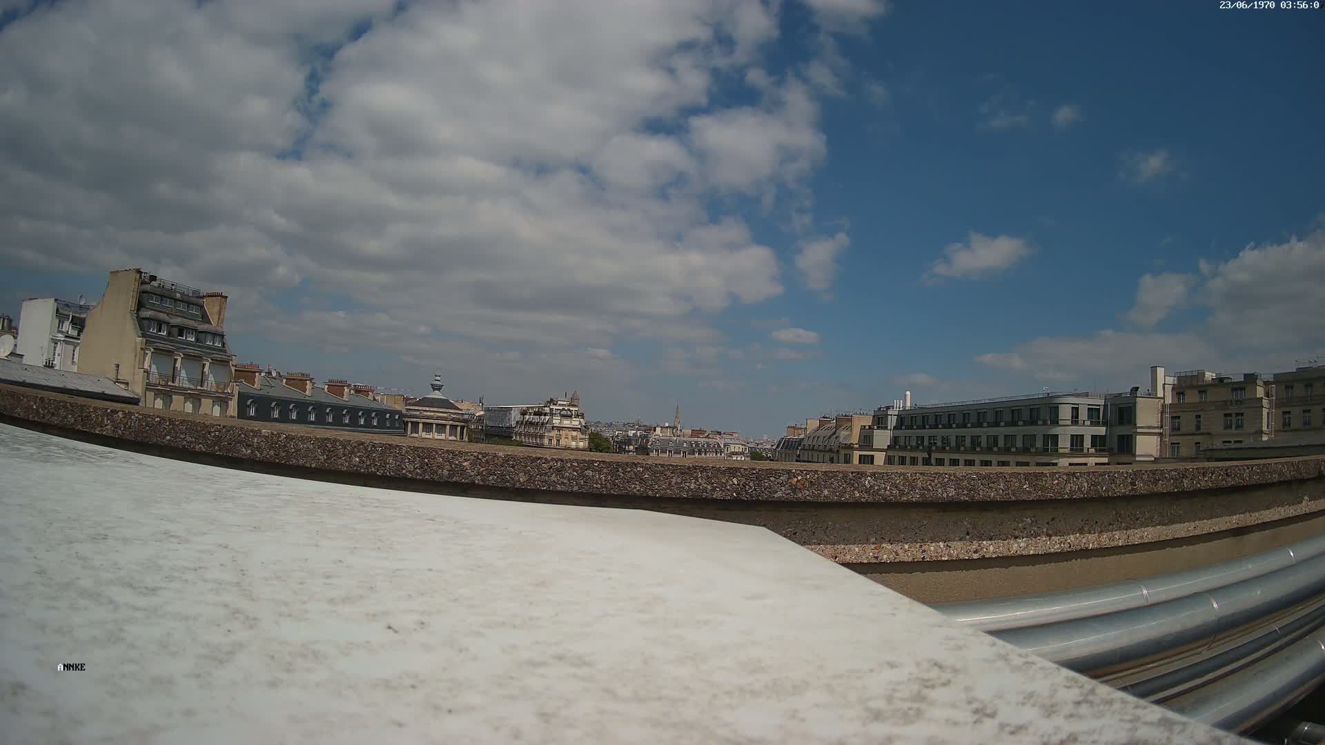 A mostly sunny day with partly cloudy skies reveals a cityscape of Parisian-style buildings viewed from a rooftop, partially obscured by a low wall and large pipes.