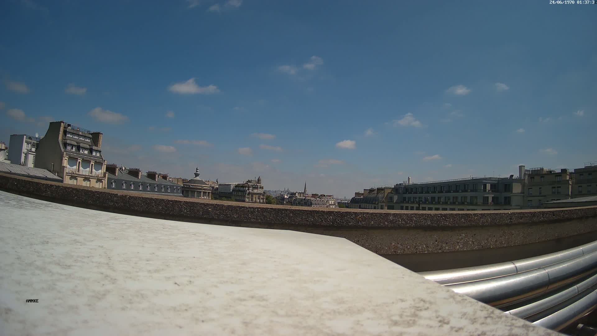 A sunny cityscape is viewed from a rooftop, with several buildings and a partly cloudy blue sky visible in the background, and large metal pipes in the foreground.