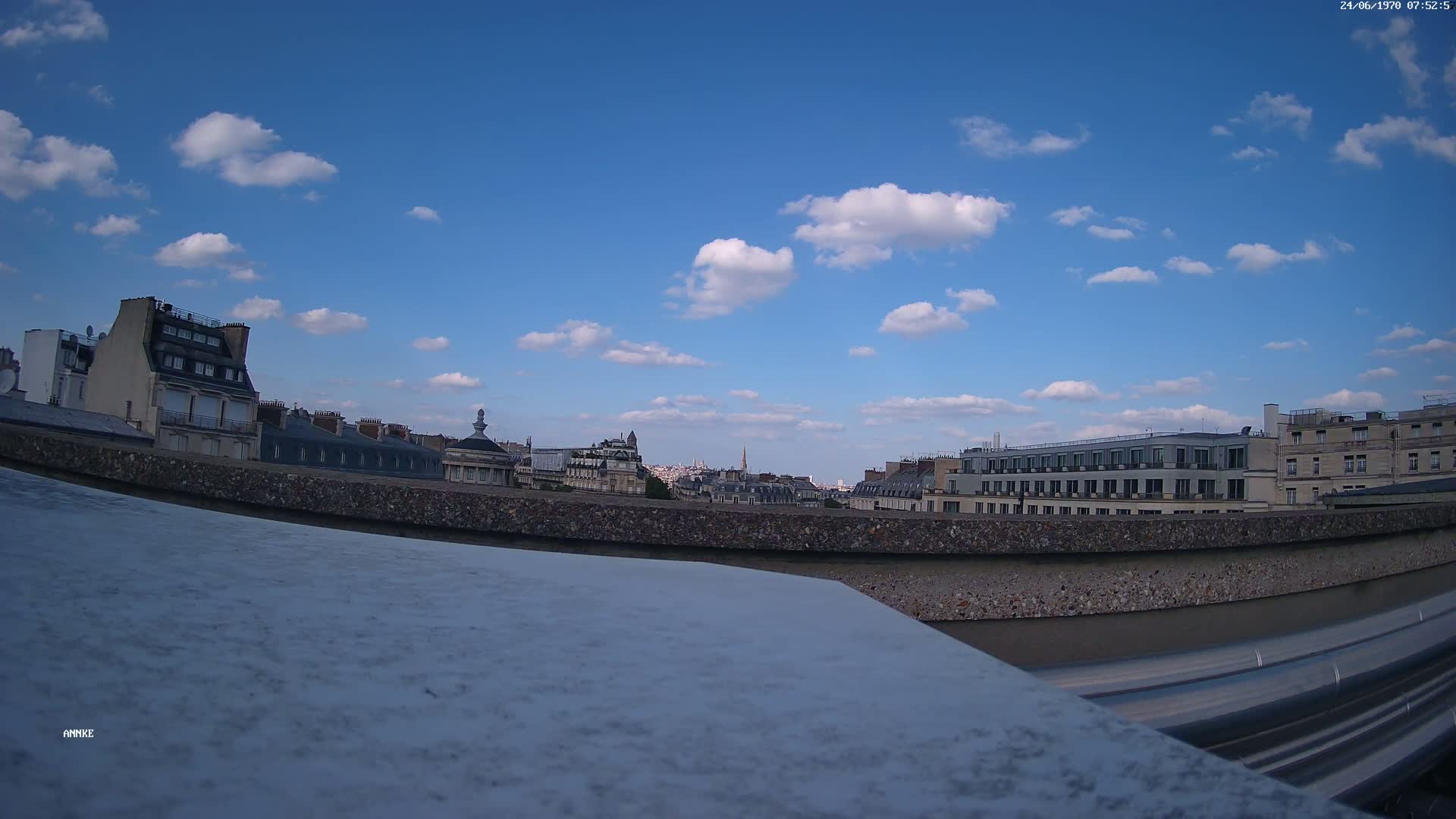 A cityscape of Parisian buildings under a mostly sunny sky with scattered cumulus clouds is seen from a rooftop vantage point.