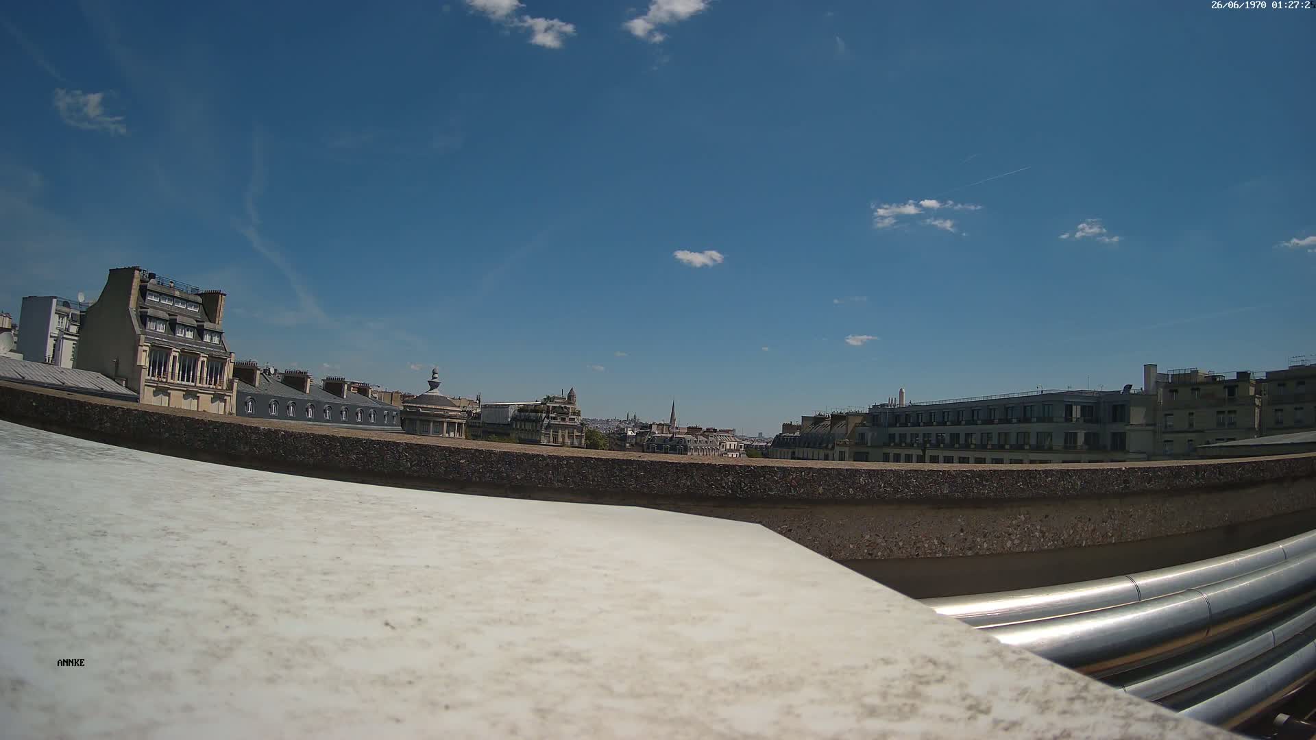 A rooftop view on a sunny day shows a cityscape of Parisian buildings, with a low wall and metal pipes in the foreground.