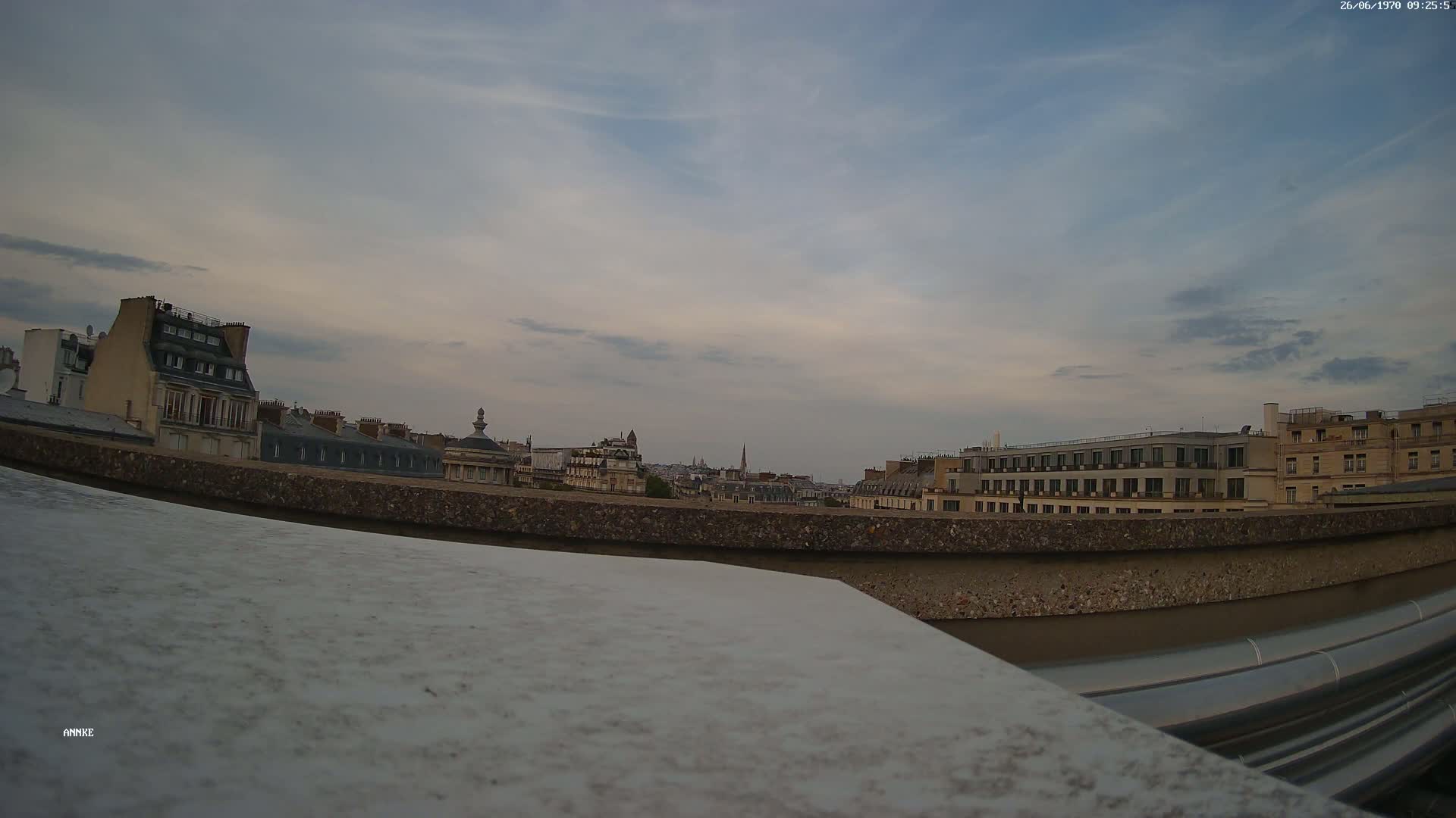 A cityscape of Parisian buildings under a partly cloudy, dusky sky is viewed from behind a low wall.
