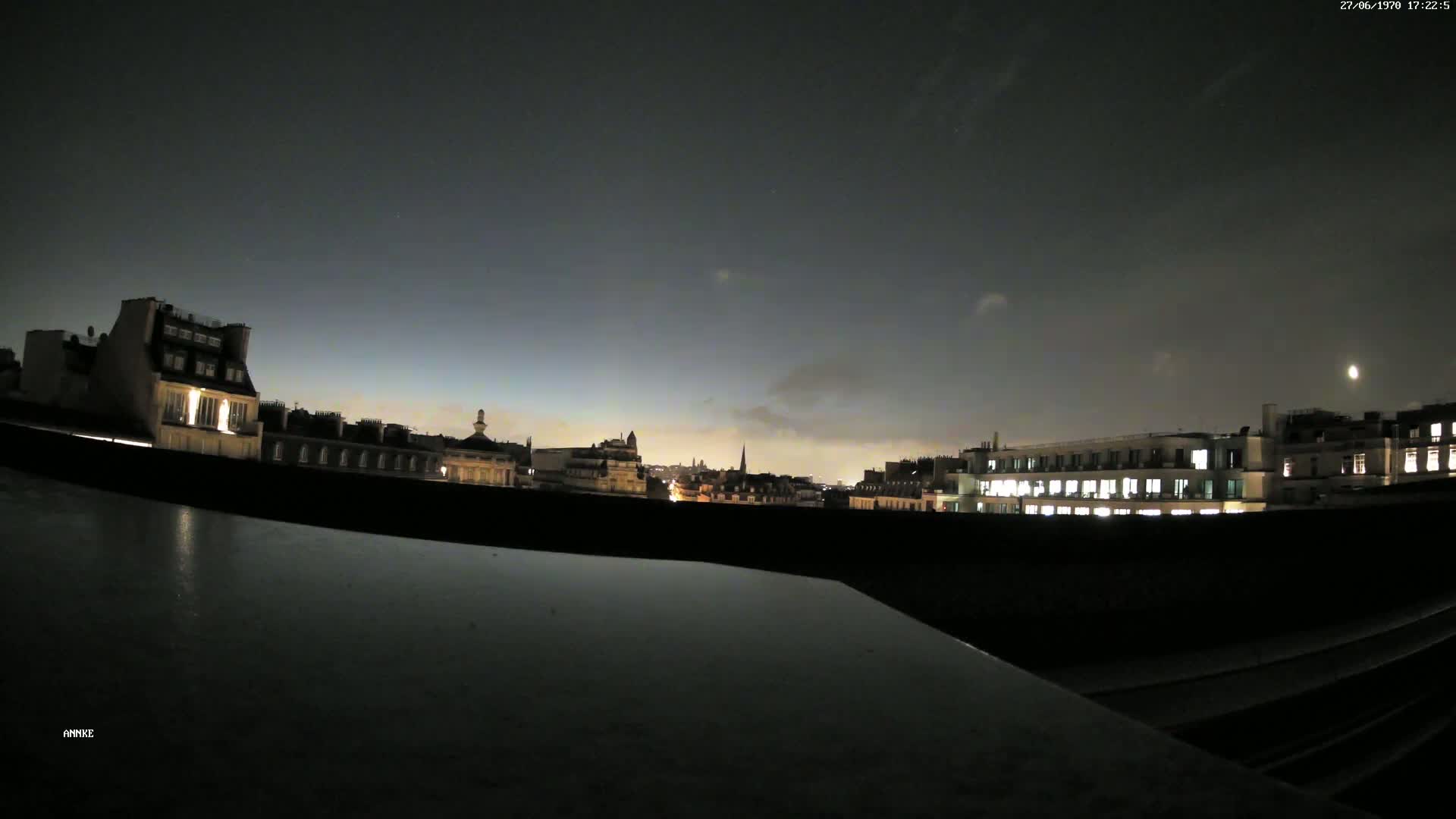 A nighttime cityscape with some clouds is viewed from a rooftop, showing city lights and a partially visible moon in the sky.