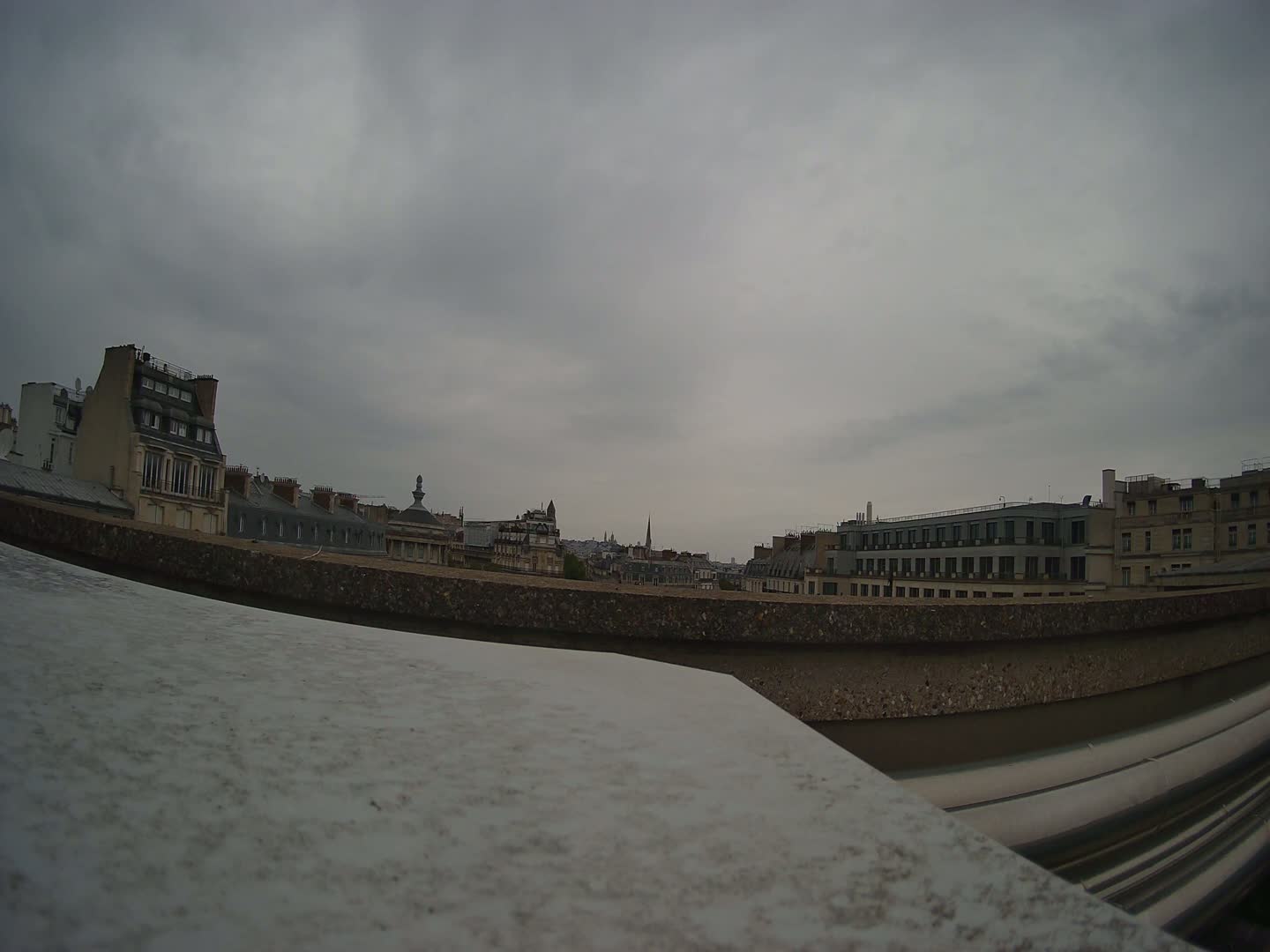 A low-angle view shows a cityscape under an overcast sky, seen from behind a low wall.