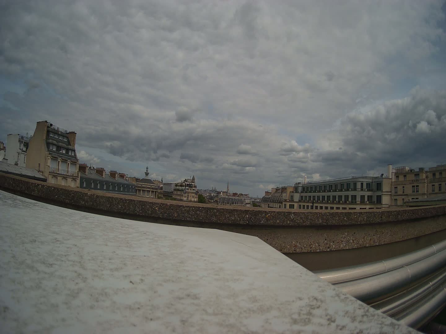 A low-angle view from a rooftop shows a cityscape under a cloudy sky, with a concrete wall and metallic pipes in the foreground.