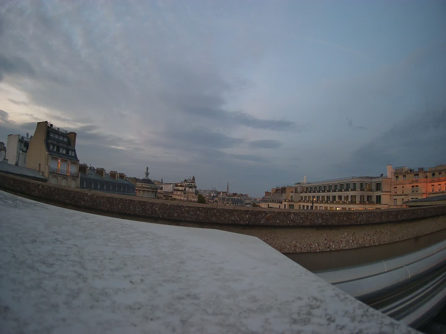 A cityscape of multi-story buildings under a cloudy, twilight sky is seen from behind a low, curved wall.
