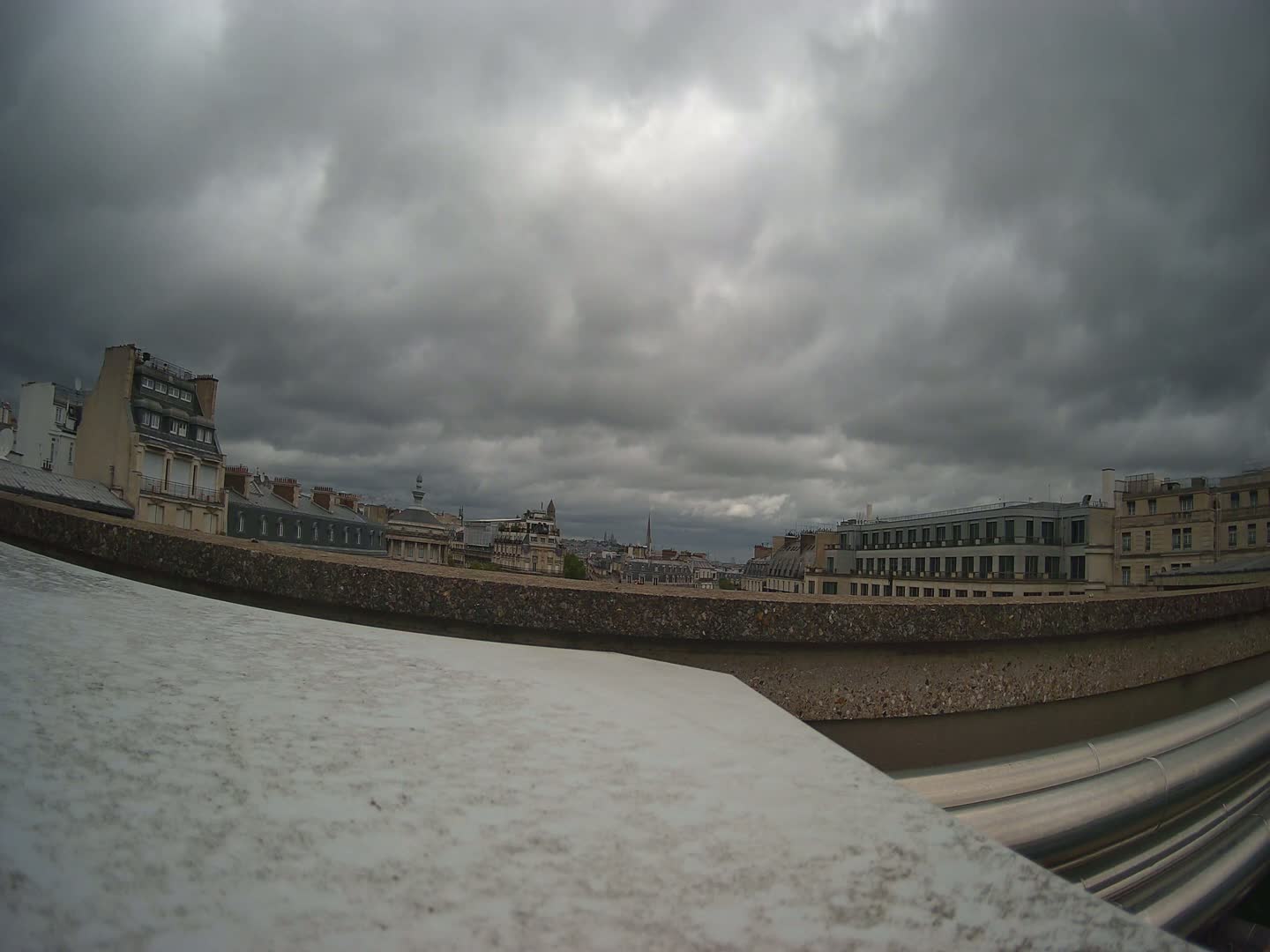 A view of a city skyline under a heavily overcast sky, seen from behind a low wall with metal piping.